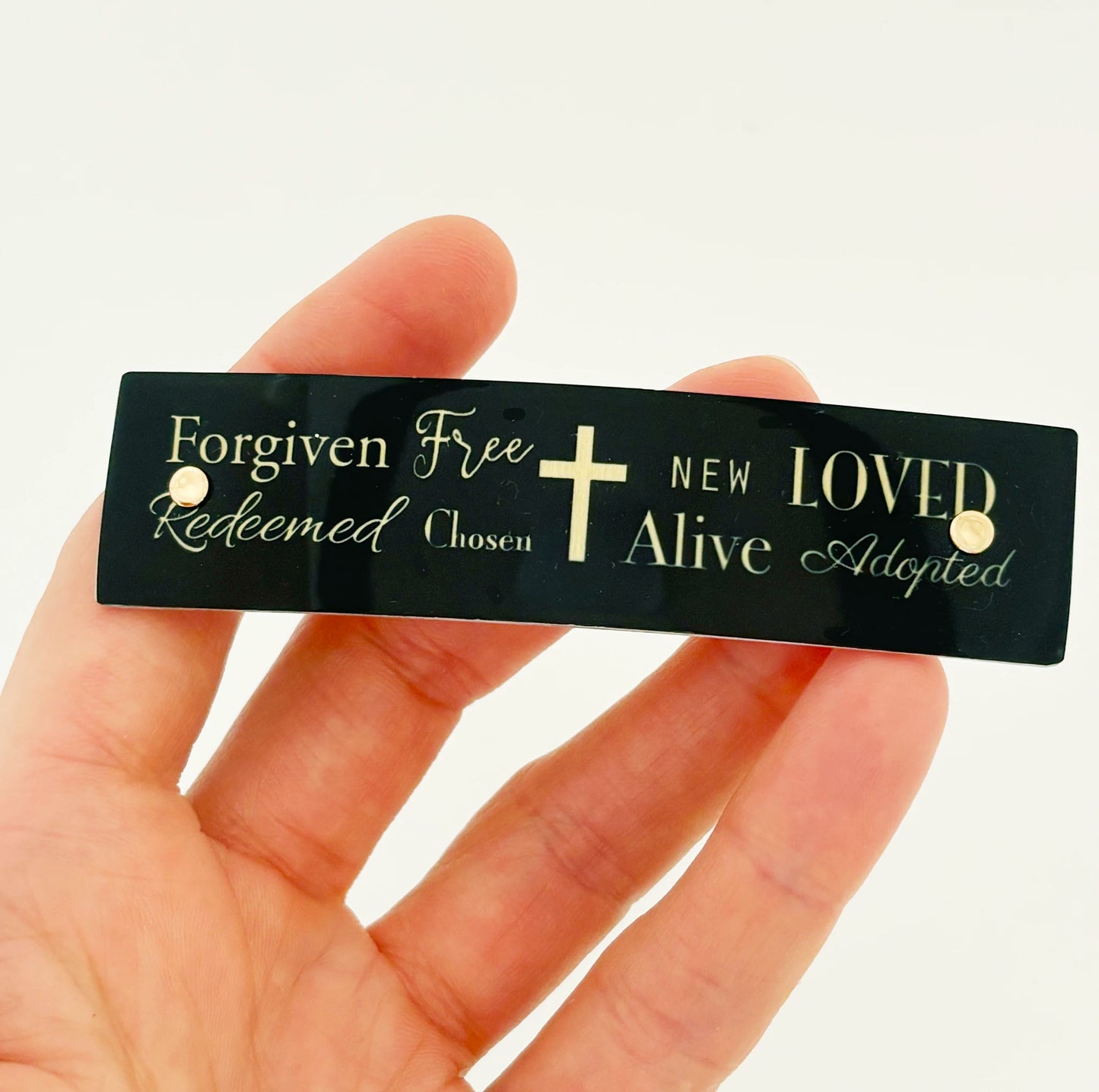 A hand holding a black rectangular hair barrette with gold text and a gold cross. The text reads 'Forgiven Free Redeemed Chosen NEW LOVED Alive Adopted' .