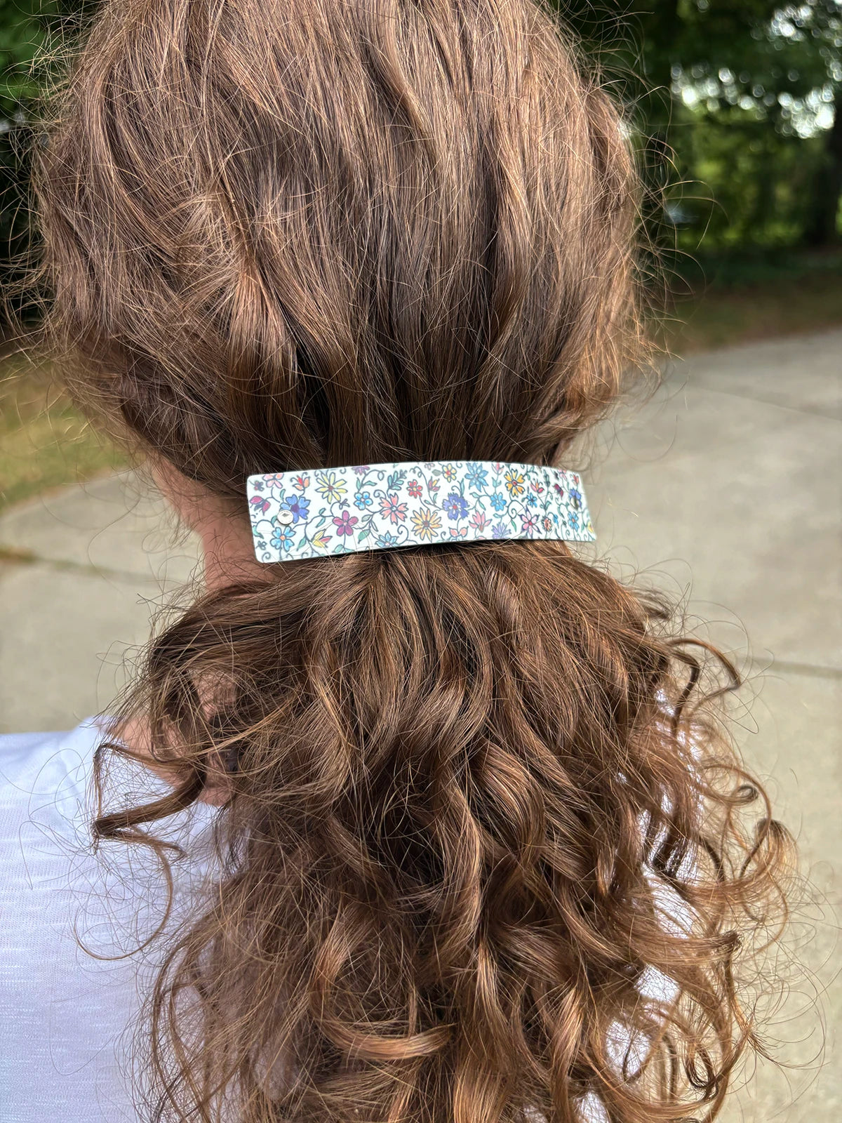A close-up back view of a person with long, wavy brown hair wearing a rectangular hair clip adorned with a colorful floral pattern on a white background.