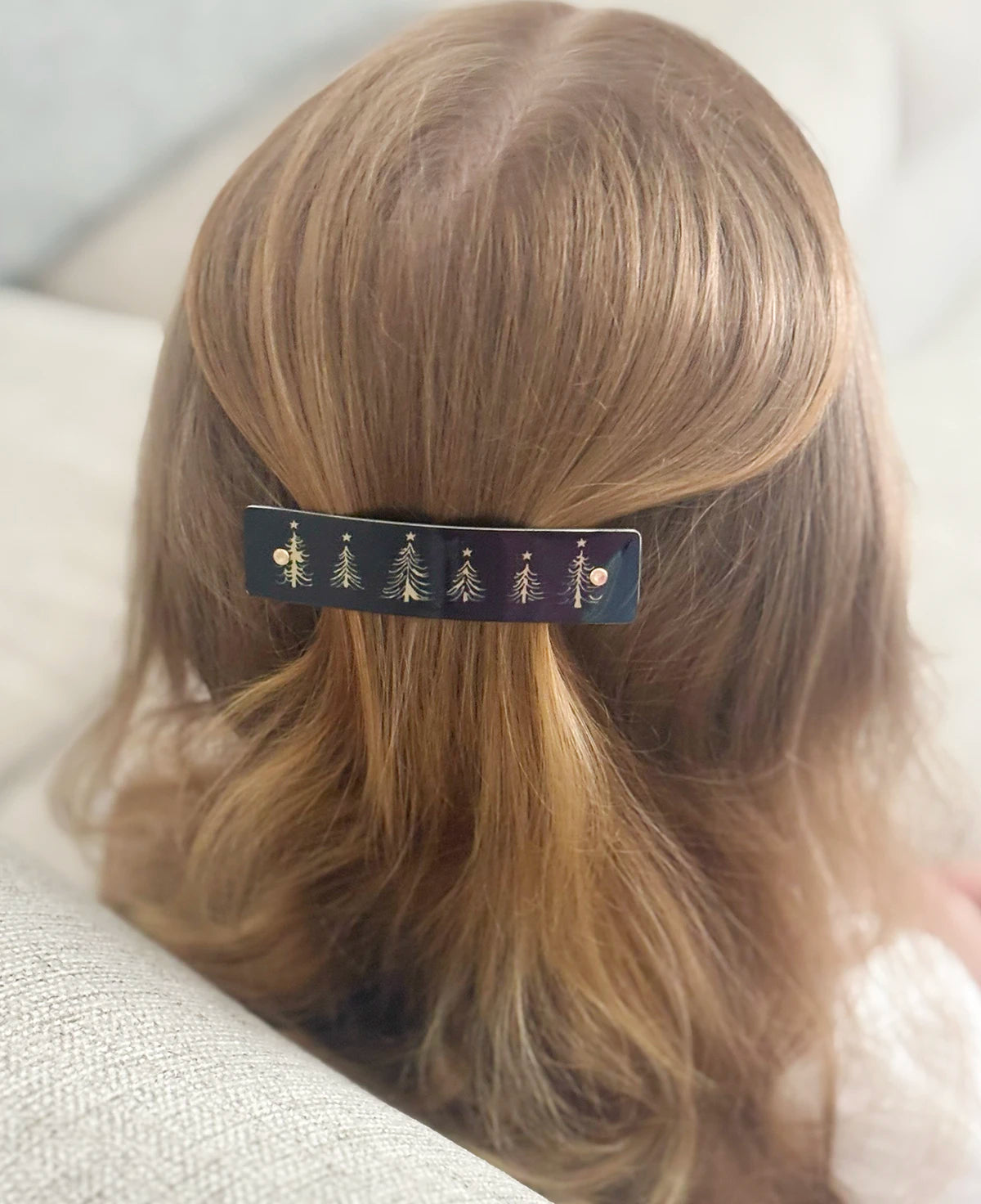 A close-up back view of a person with light brown hair wearing a black hair clip decorated with small gold Christmas tree and star motifs.
