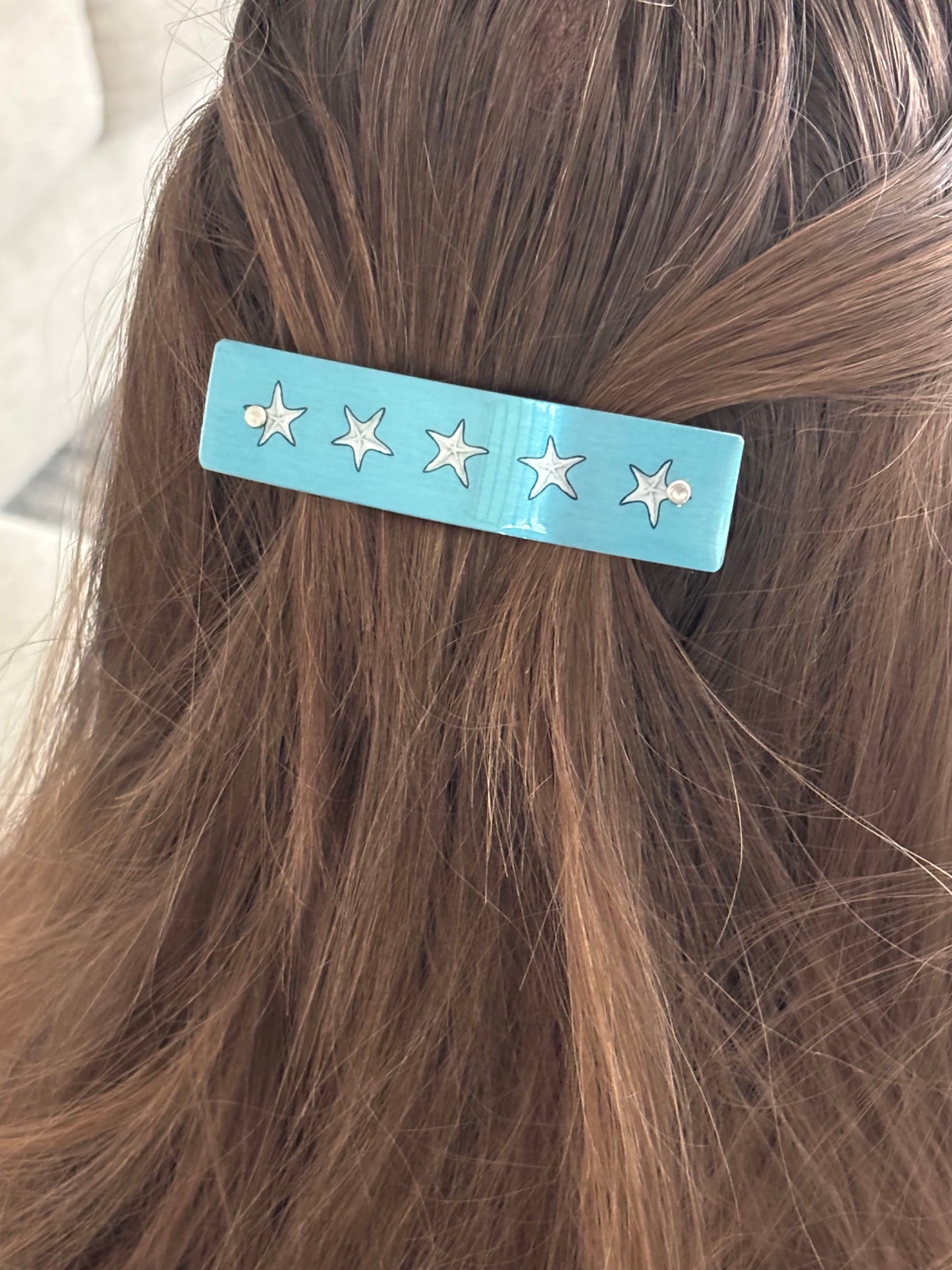 A light blue hair clip with five white starfish designs, shown clipped to a section of brown hair.