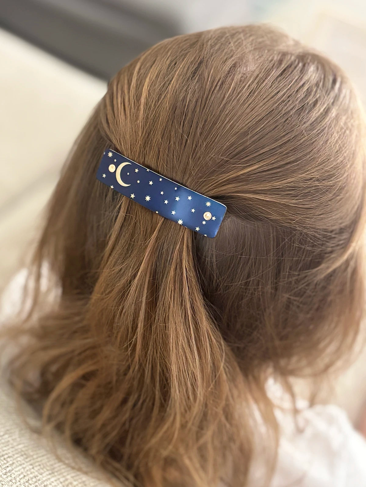 A close-up of a child's light brown hair secured with a blue hair clip decorated with a gold crescent moon and stars, shown from behind.
