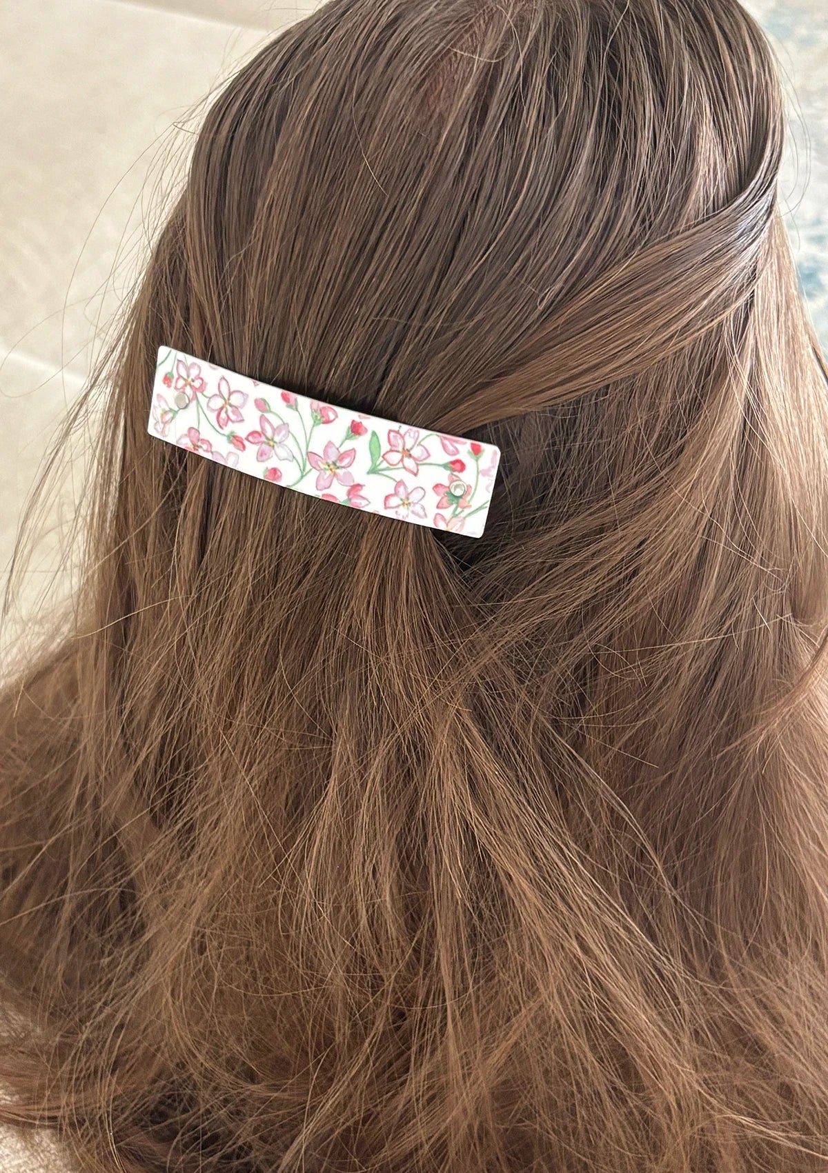 A close-up of a person's brown hair secured with a white handmade boutique barrette hair clip decorated with an original watercolor pink and green apple blossom pattern.
