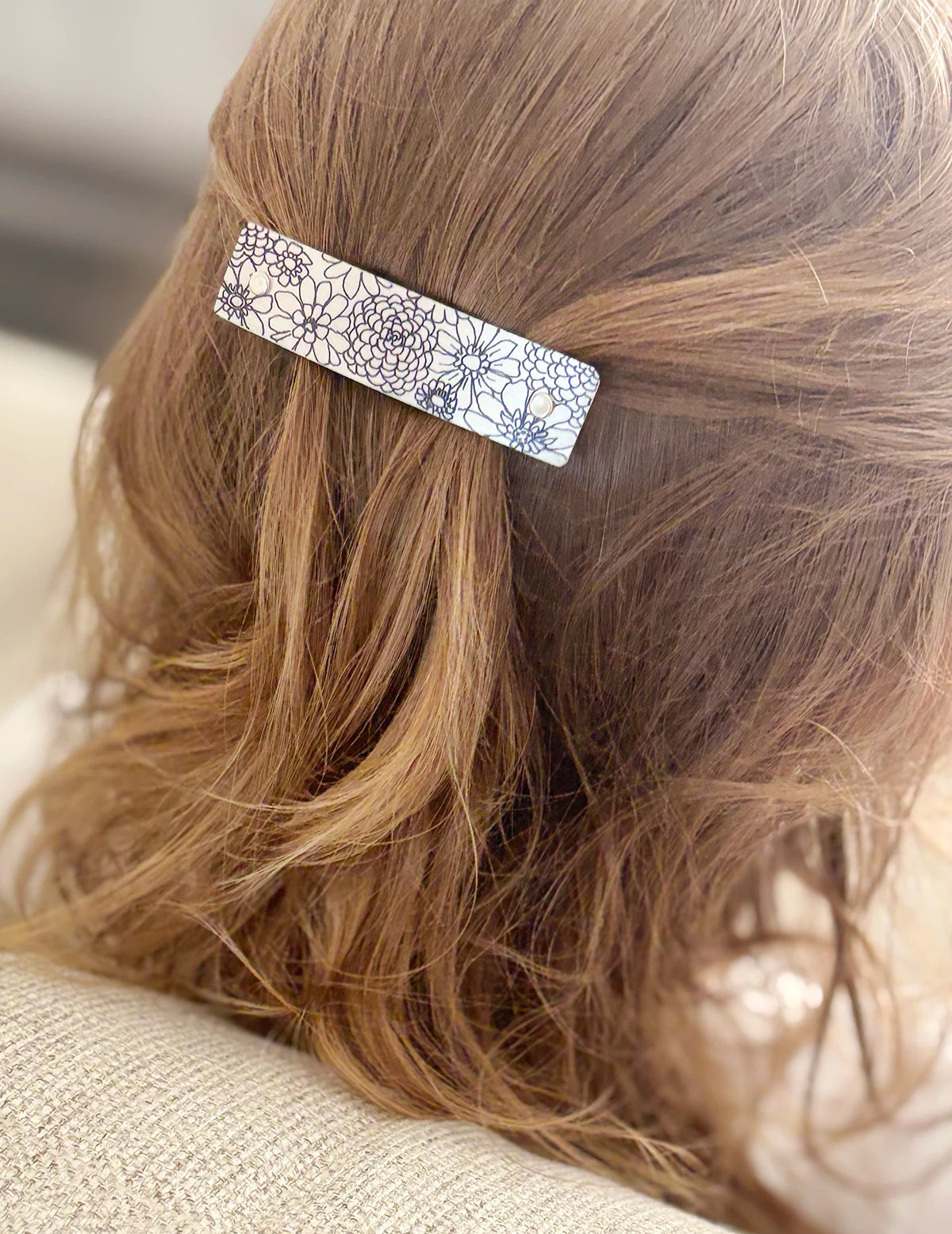A close-up of a person's light brown hair secured with a slim rectangular boutique hair clip featuring original artwork of a black floral pattern on a silver background.