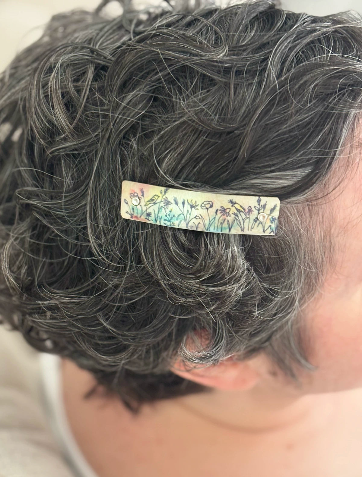 A close-up of a person's short, curly gray hair wearing a small rectangular hair clip decorated with a colorful wildflower pattern.