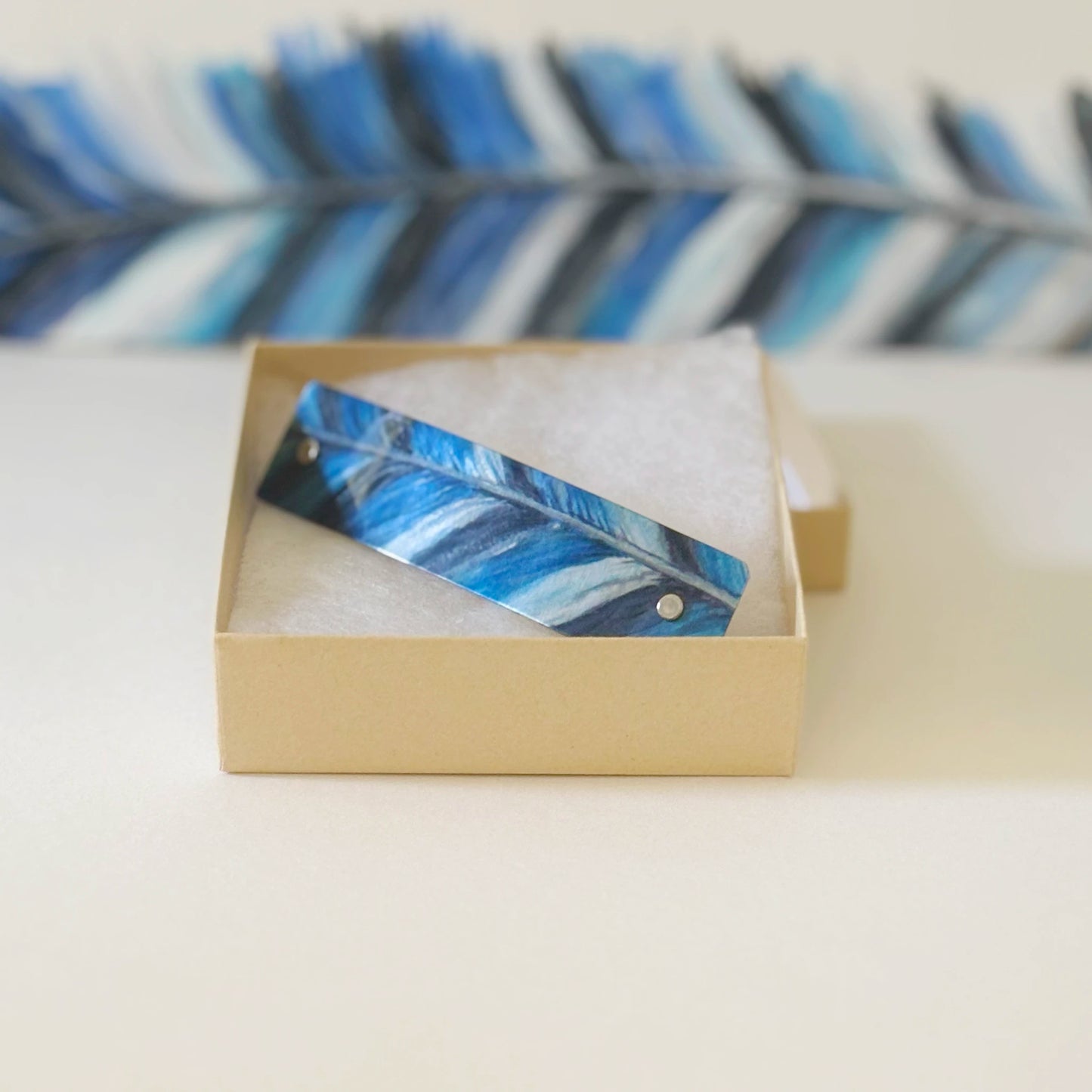 A rectangular feather French barrette with a blue, white, and black feather pattern rests inside an open beige kraft paper gift box on a light cream surface, with a blurred matching handpainted feather in the background.