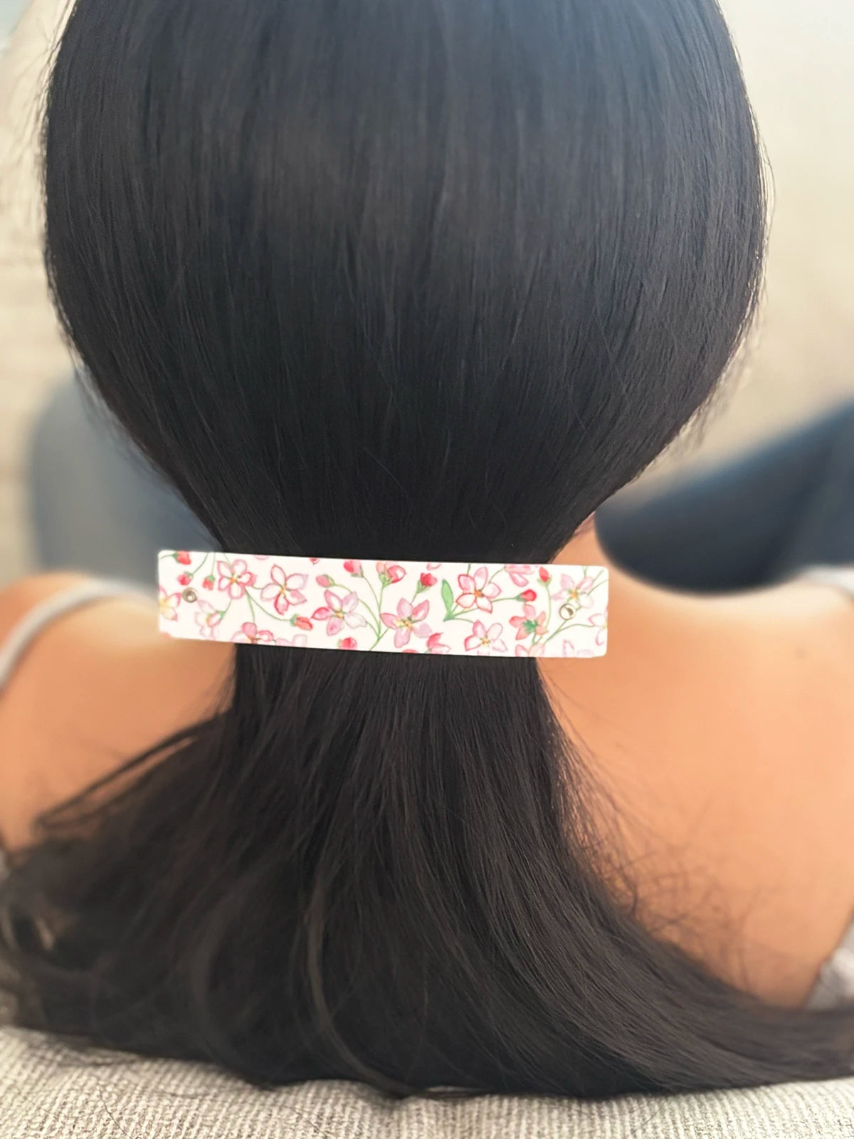 A close-up back view of a person with long black hair secured in a low ponytail, wearing a slim rectangular handmade hair clip with a white background and a pink and green floral pattern.