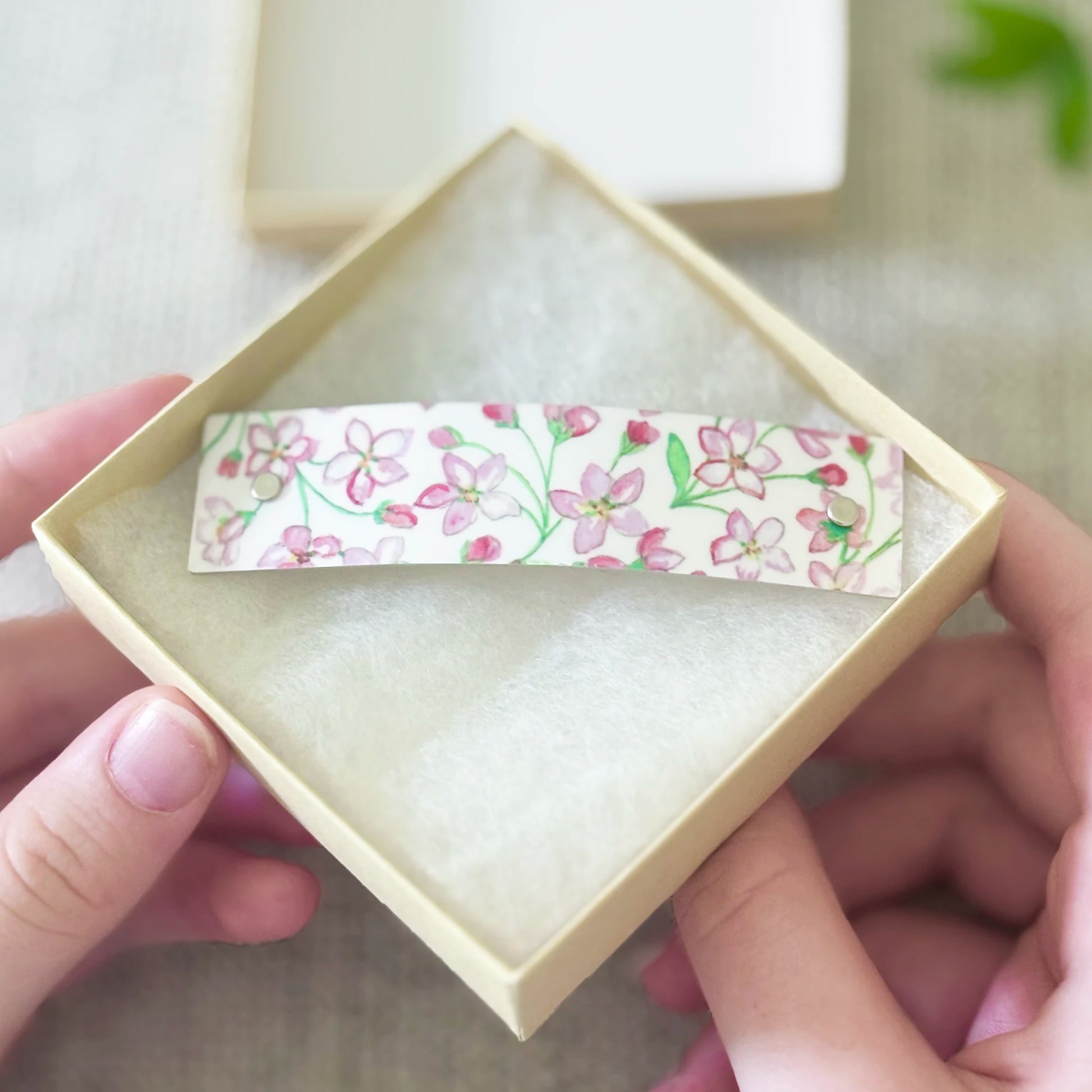 A close-up of hands holding an open beige gift box with a small rectangular white hair clip barrette featuring an artistic watercolor pink floral pattern, set against a soft neutral background.