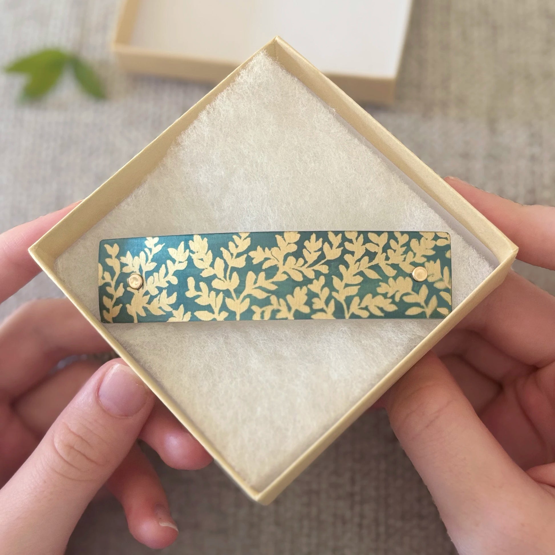 A rectangular barrette with a dainty gold leaf pattern on a dark teal-green background, held in a cream-colored square gift box with a soft cream insert. Two hands are holding the box, and a soft-focus background is visible.