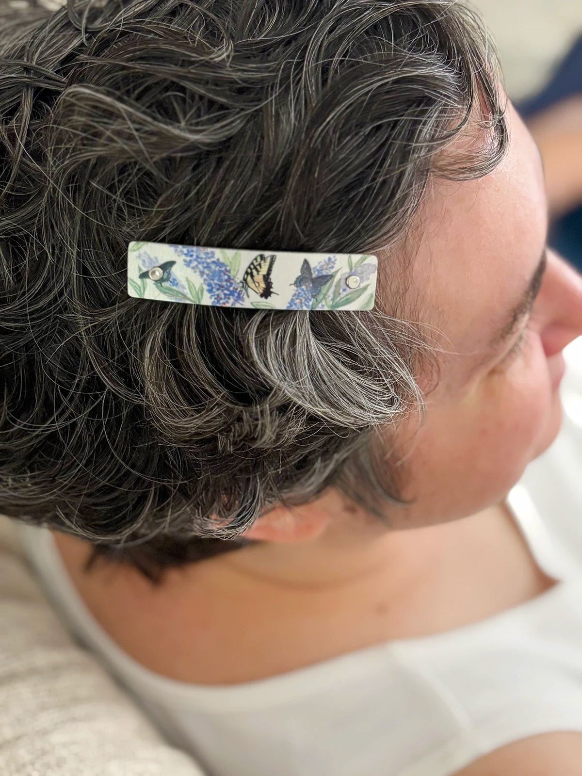 A close-up side profile of a person with short, dark gray hair wearing a rectangular hair clip adorned with painted butterflies and blue flowers.
