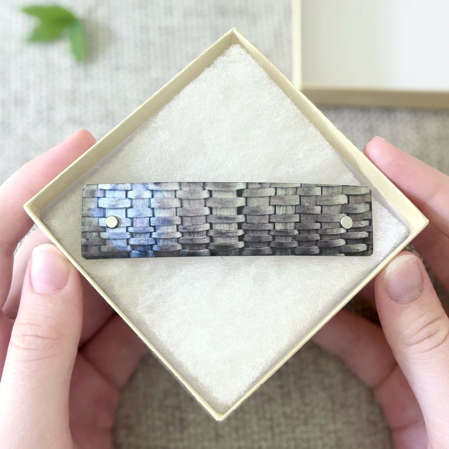 A rectangular woven hair barrette with a textured gray and black pattern, presented inside a cream-colored square gift box held by two hands.