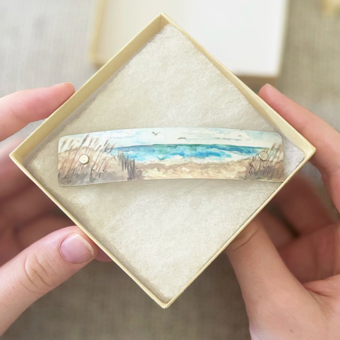 A handcrafted artistic hair barrette clip with a painted beach scene by Robbin V. Davis, held in a diamond-shaped presentation box by two hands.