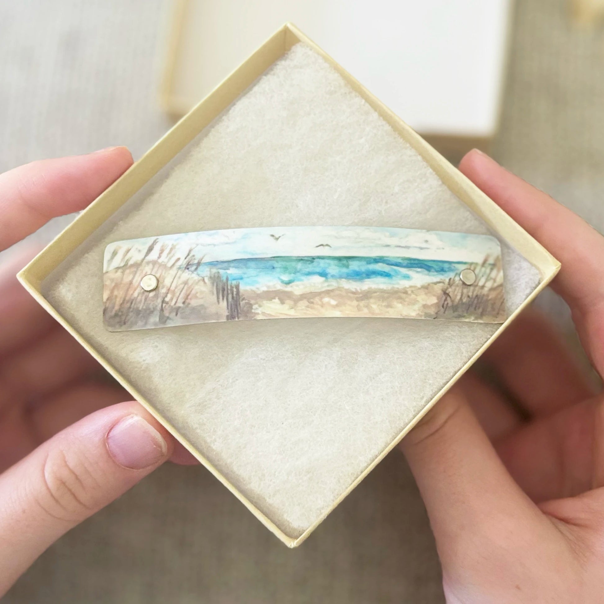 A handcrafted artistic hair barrette clip with a painted beach scene by Robbin V. Davis, held in a diamond-shaped presentation box by two hands.