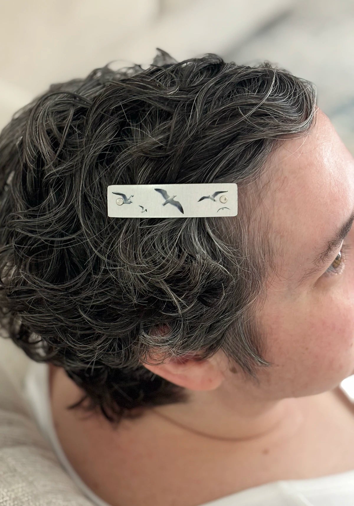 A close-up of a person's short, curly gray hair wearing a silver hair clip decorated with shorebirds.