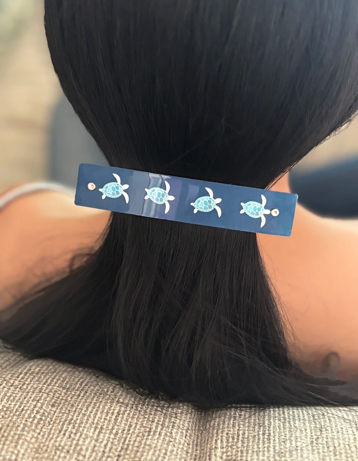 A close-up of a person's dark hair tied back in a low ponytail, featuring a navy blue rectangular hair clip decorated with small sea turtle illustrations.