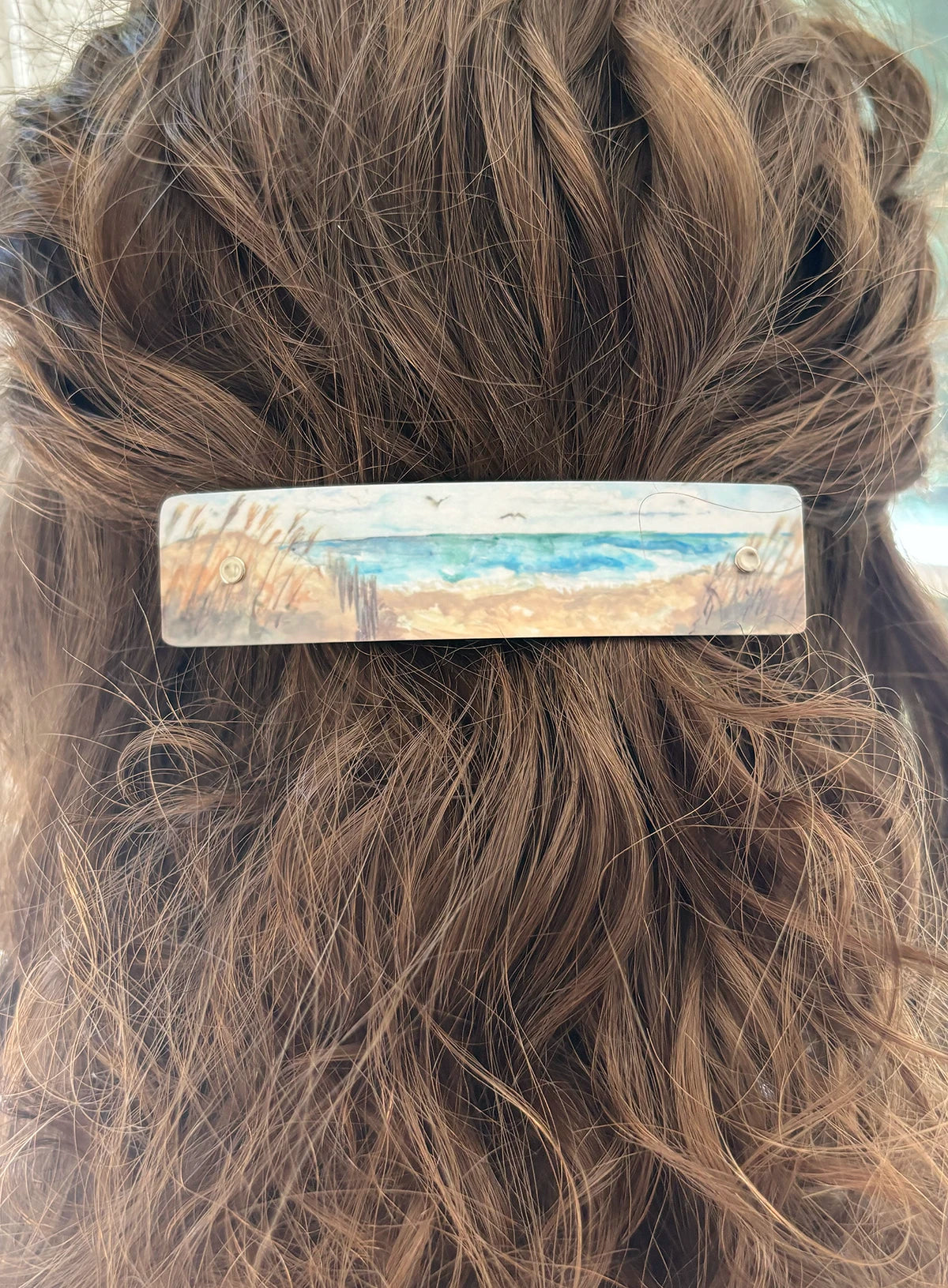 A close-up of a person's brown hair with a rectangular barrette hair clip featuring a painted beach landscape design in blue, beige, and brown tones, handmade by Robbin V. Davis Studio in North Carolina.