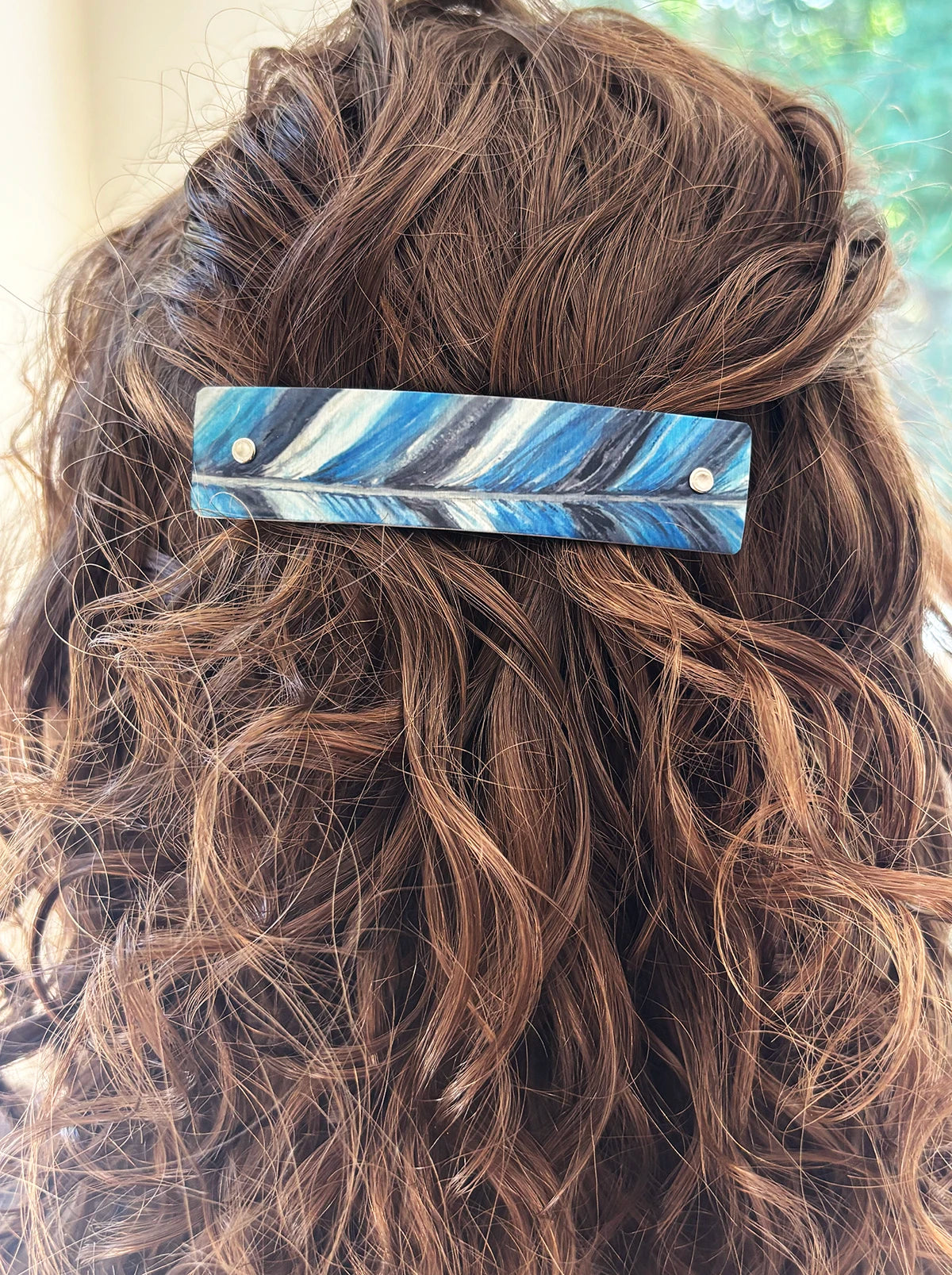 A close-up of a person's wavy brown hair holding a rectangular hair clip with a blue, white, and black feather-like pattern.