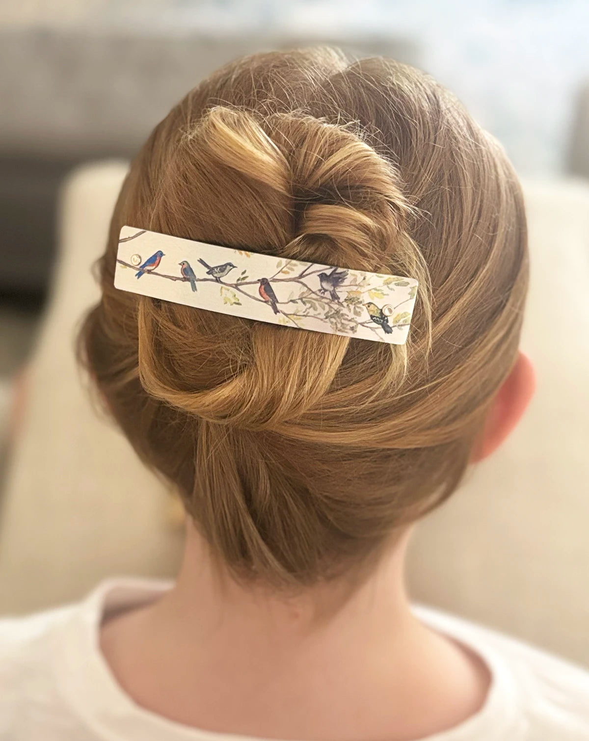 A close-up back view of a person wearing a light silver barrette hair clip decorated with a delicate bird and branch pattern, holding up a styled blonde updo.