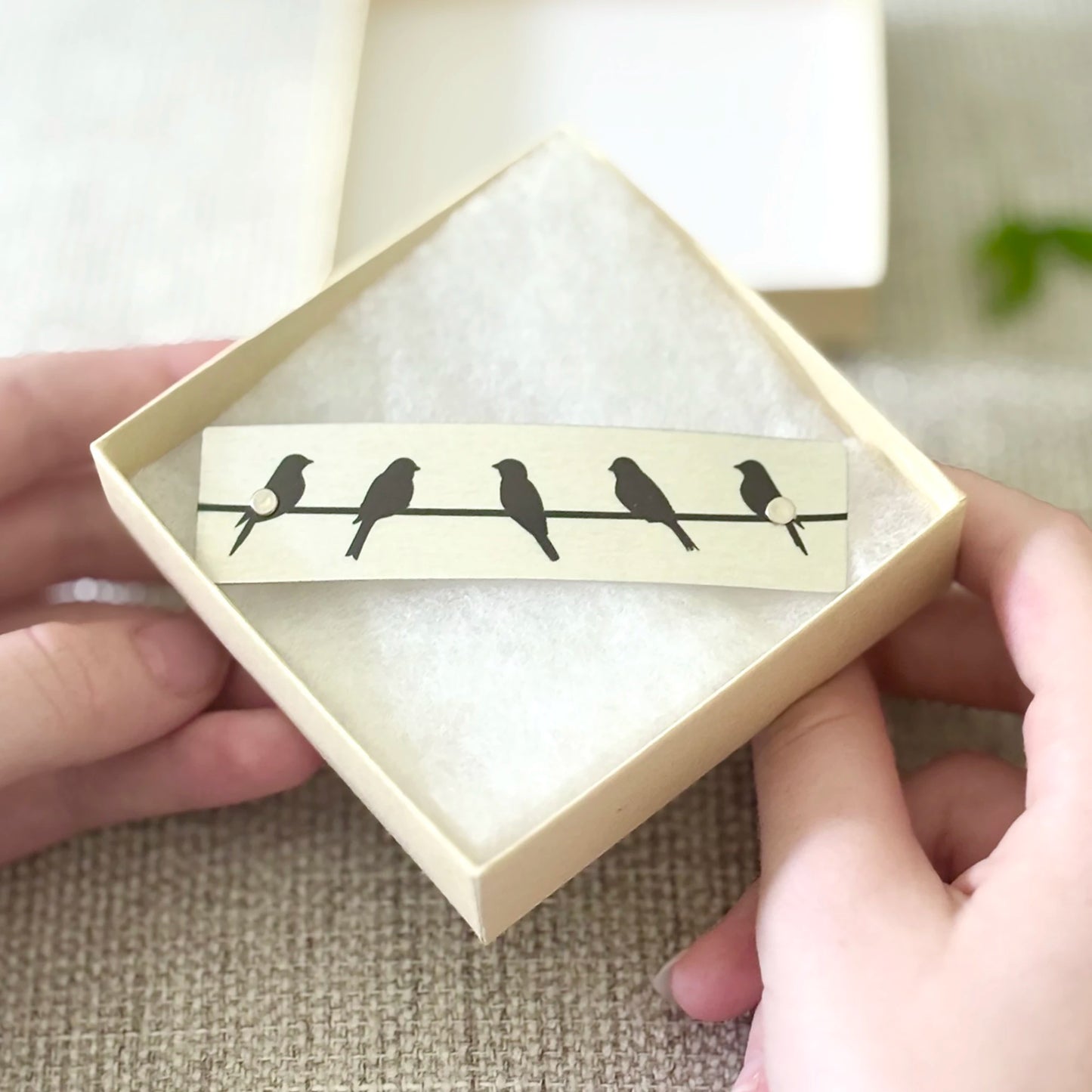 A rectangular silver barrette with black bird silhouettes perched on a thin horizontal line, presented in a small cream gift box and held by two hands against a soft neutral background.