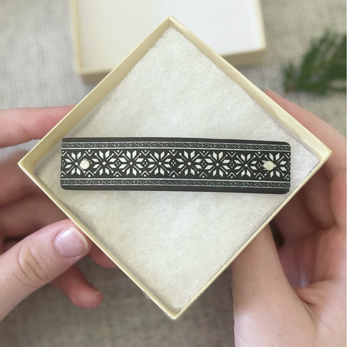 A black metal barrette with intricate silver floral cutout patterns, held in a square cream-colored presentation box with a soft white insert, held by two hands.