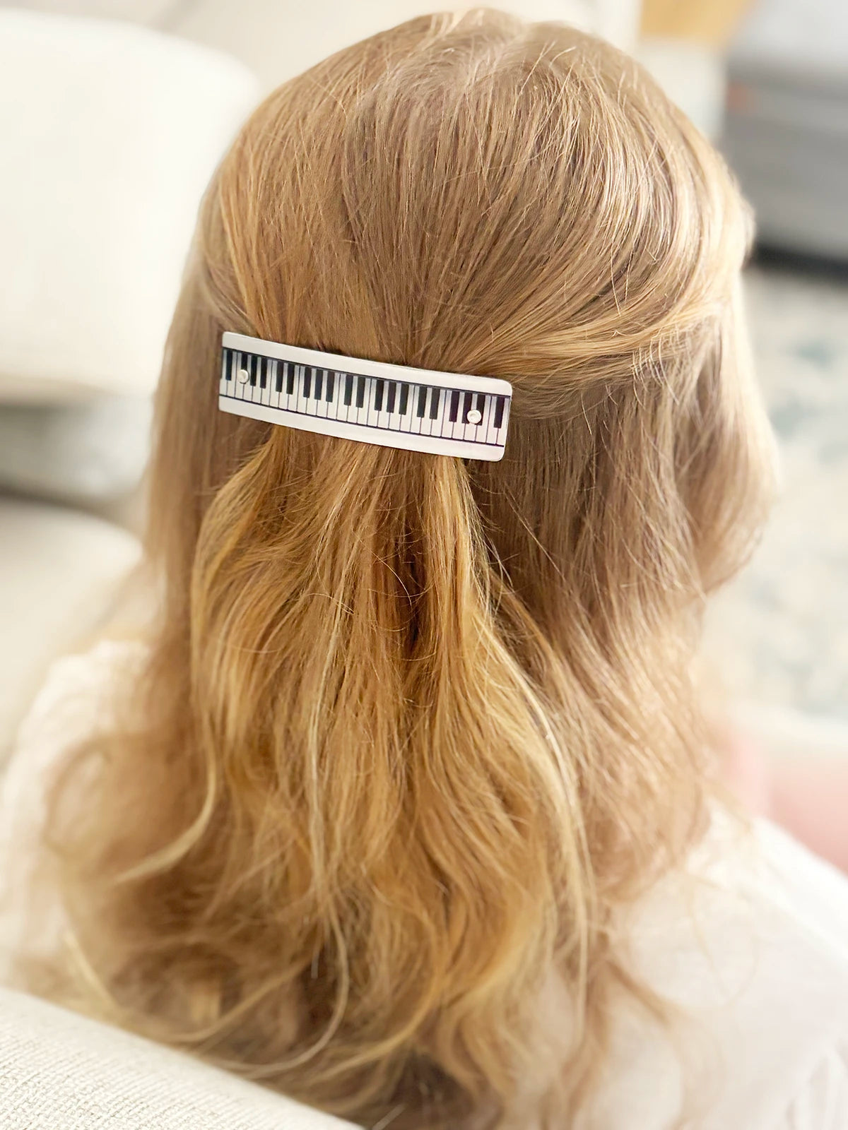 A close-up back view of a person with long blonde wavy hair, holding their hair back with a silver hair clip designed to look like a piano keyboard.