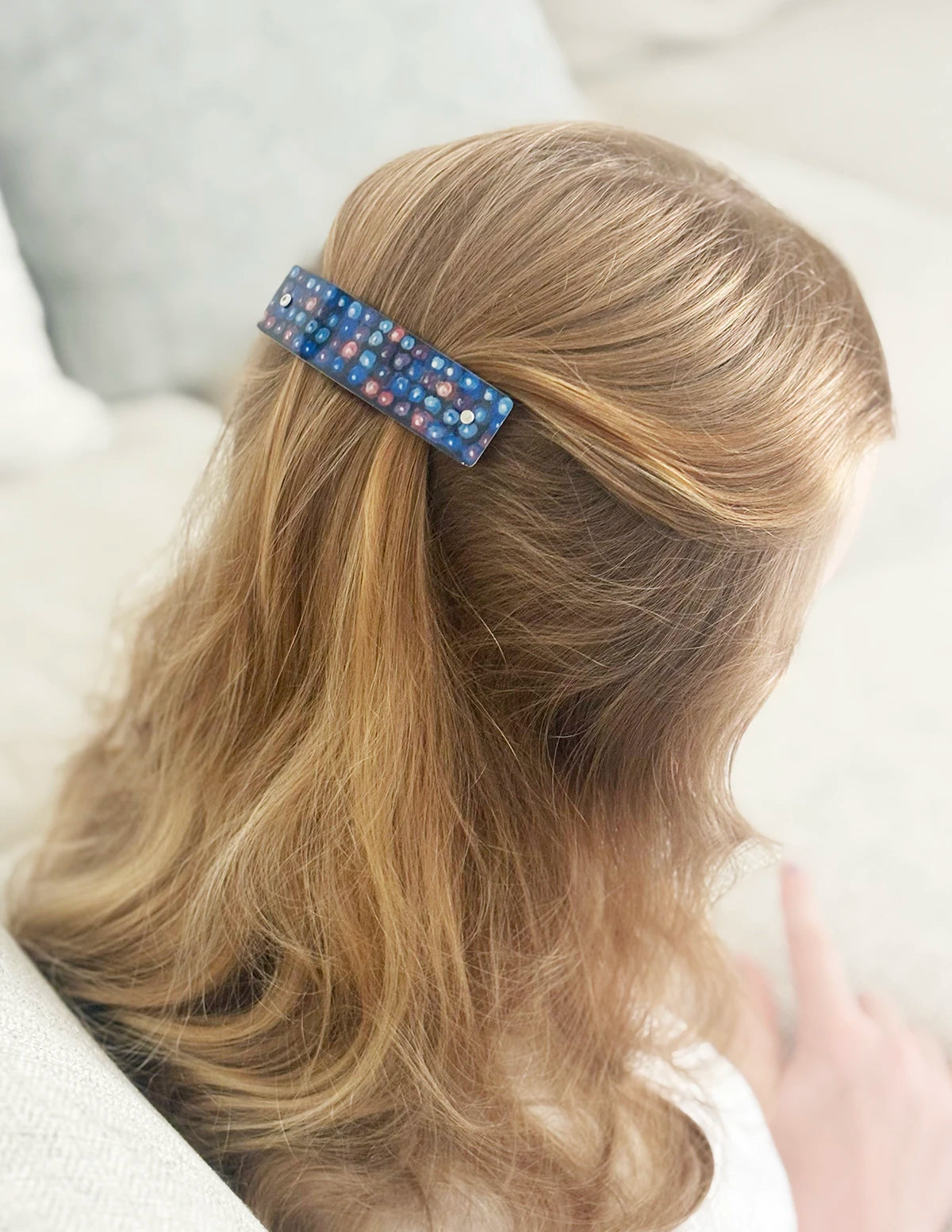 A close-up lifestyle shot of a person with blonde hair seen from behind, wearing a blue blackberry pattern  hair clip to section her hair, set against a soft, light-colored indoor background.