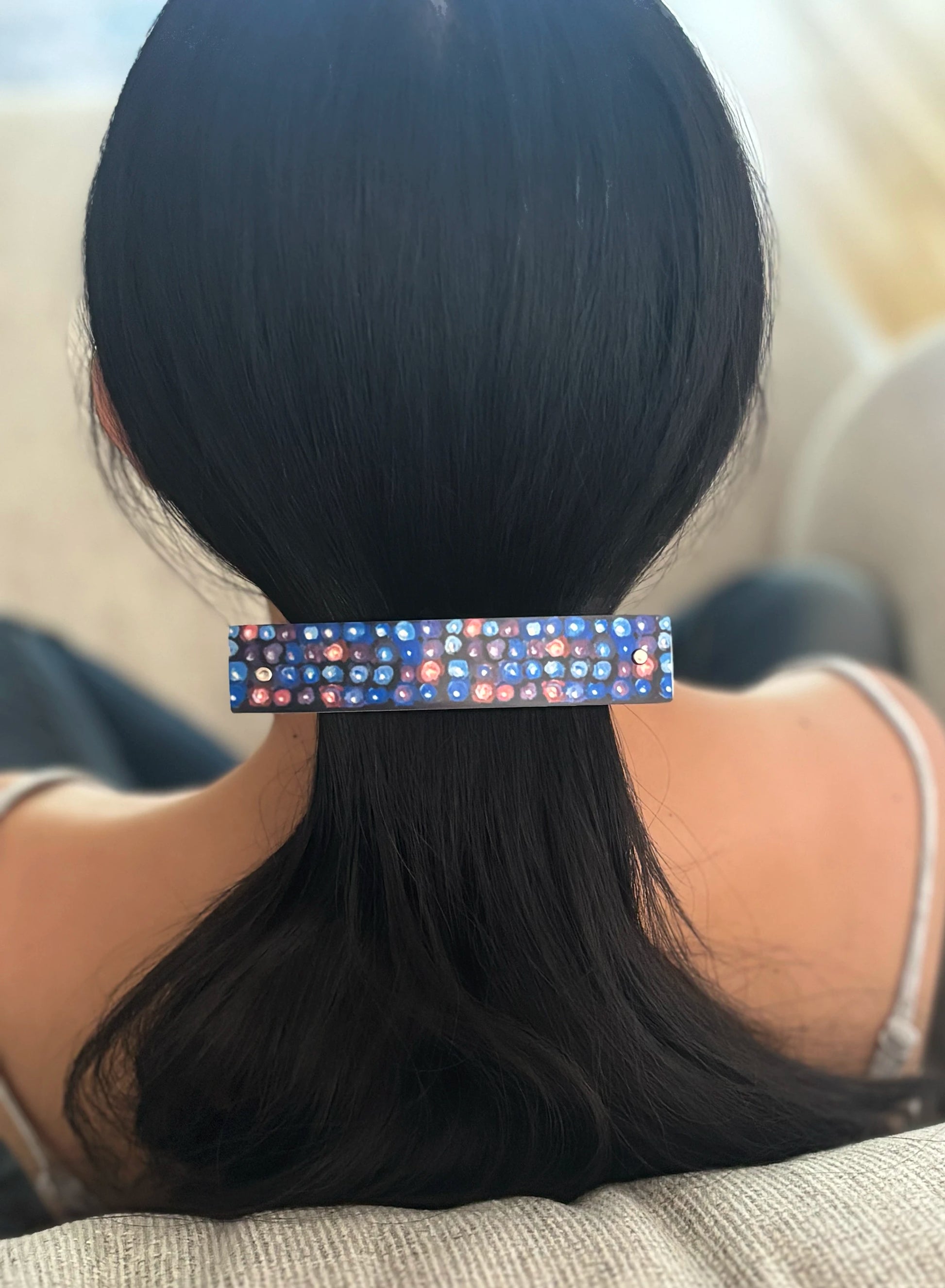A close-up back view of a person with long black hair secured by a slim rectangular hair clip decorated with a blackberry inspired design on a dark blue background.