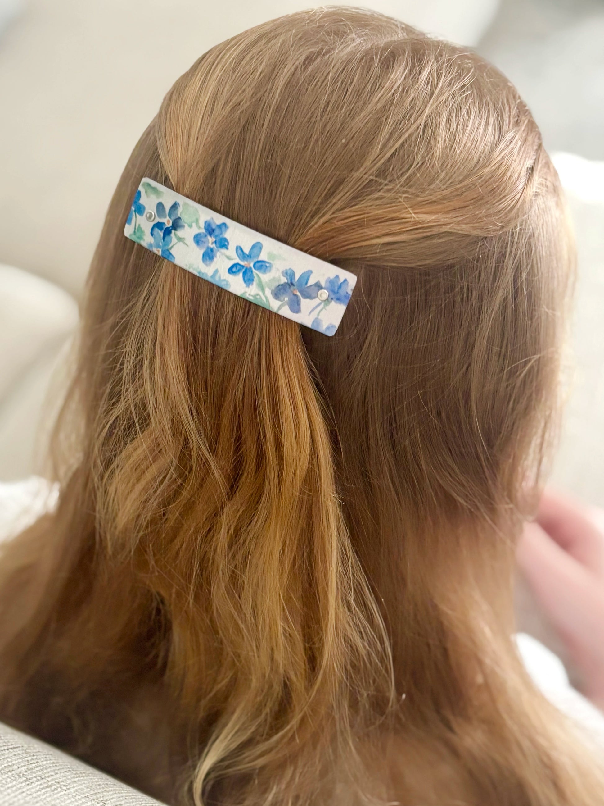 A close-up lifestyle shot of a person's light brown hair secured with a silver rectangular hair clip decorated with a blue floral pattern, set against a soft, neutral indoor background.