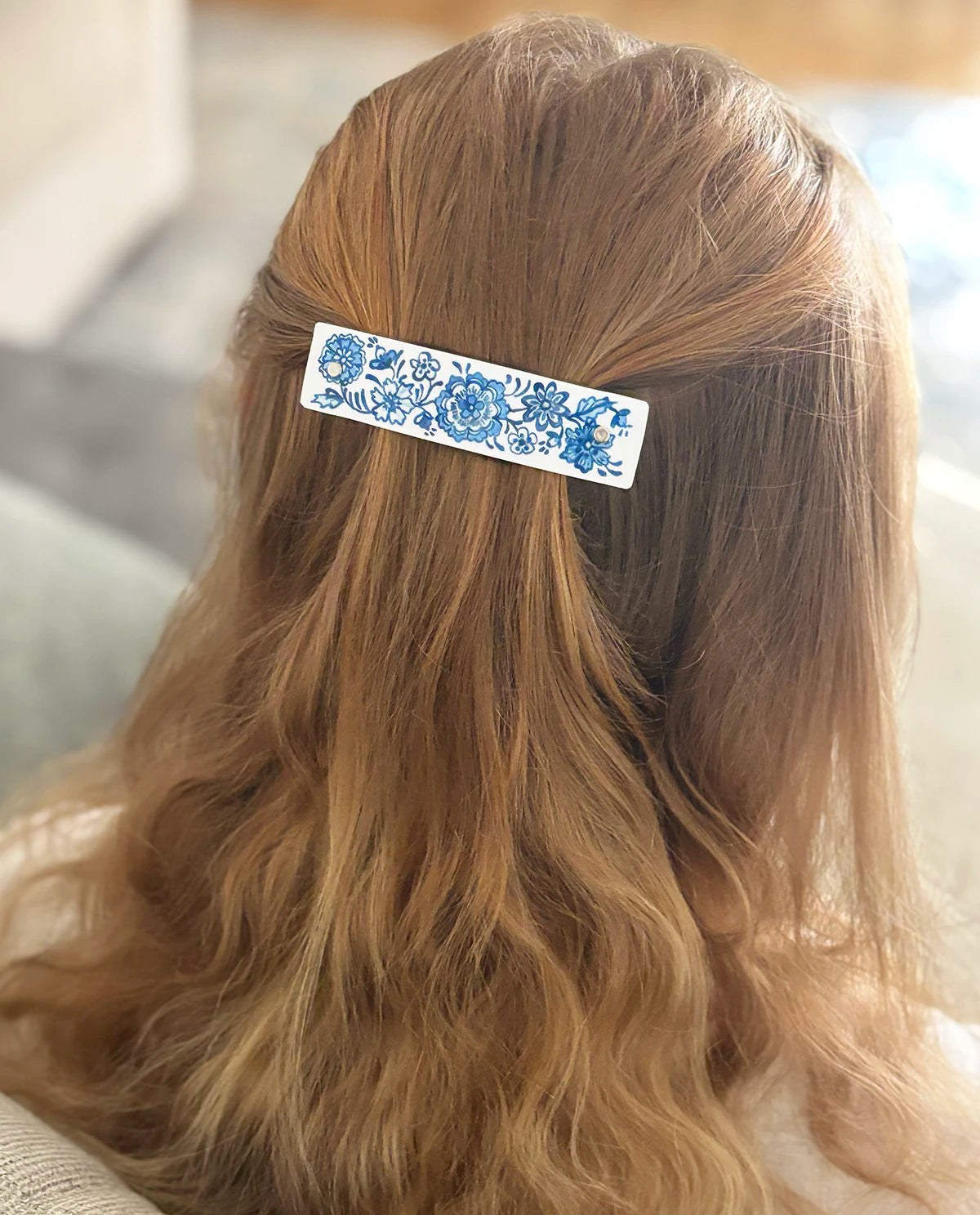 A close-up of a person's long, wavy strawberry blond hair from behind, featuring a white hair clip with a blue floral Delft pattern.