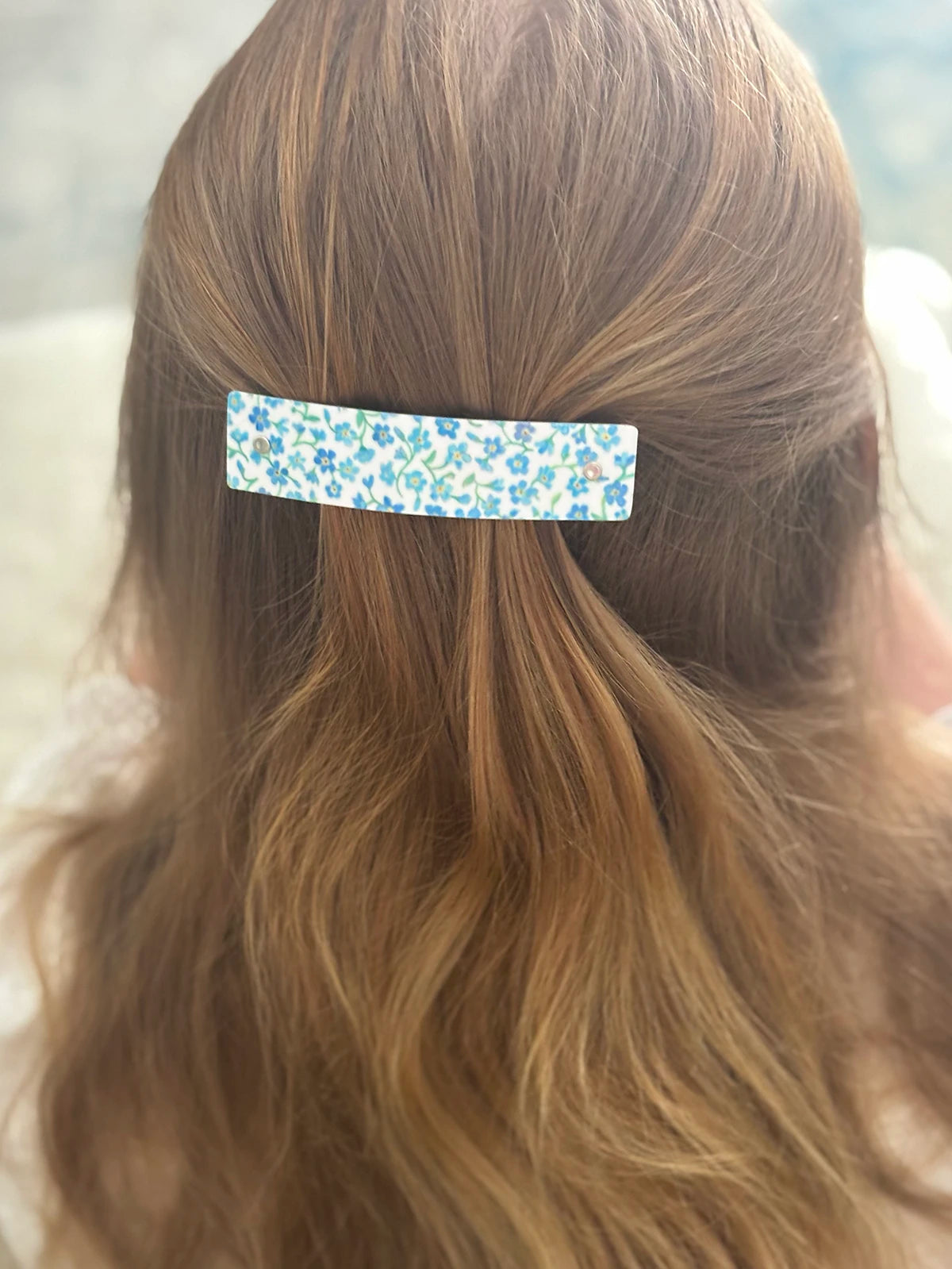 A close-up back view of a person with long light brown hair, wearing a small rectangular hair clip with a blue and white floral pattern.