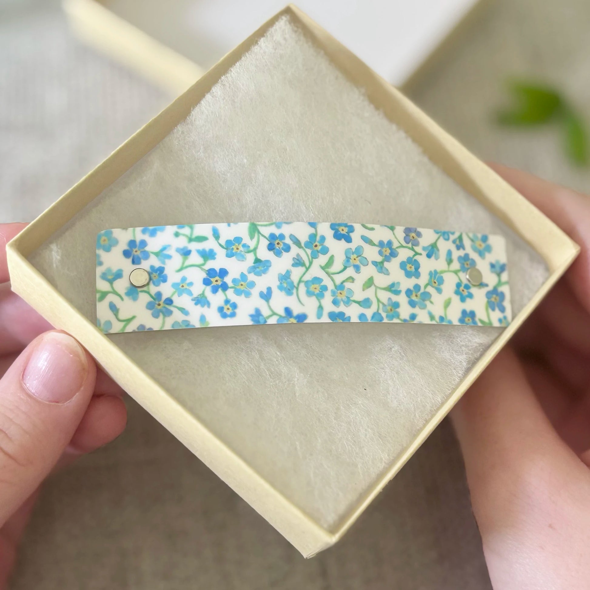 A floral-patterned hair clip held in a hand over a small cream-colored presentation box. The clip has a white background with blue and green forget-me-not flowers.
