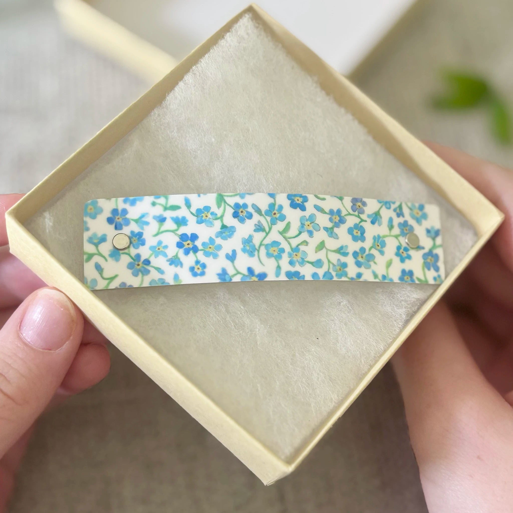 A floral-patterned hair clip held in a hand over a small cream-colored presentation box. The clip has a white background with blue and green forget-me-not flowers.
