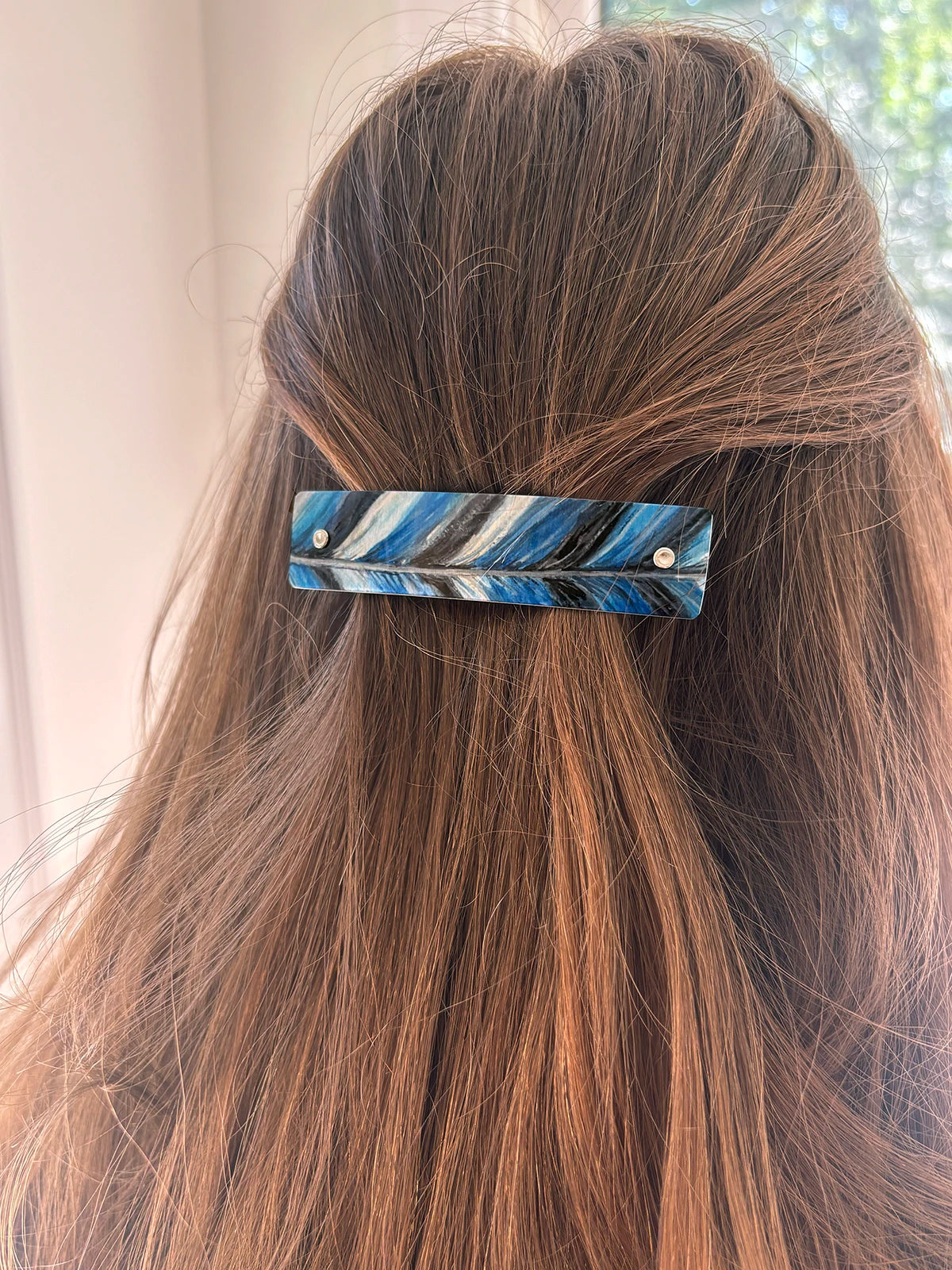 A close-up of a person's brown hair secured with a rectangular silver metal hair clip featuring a blue, black, and white feather pattern.