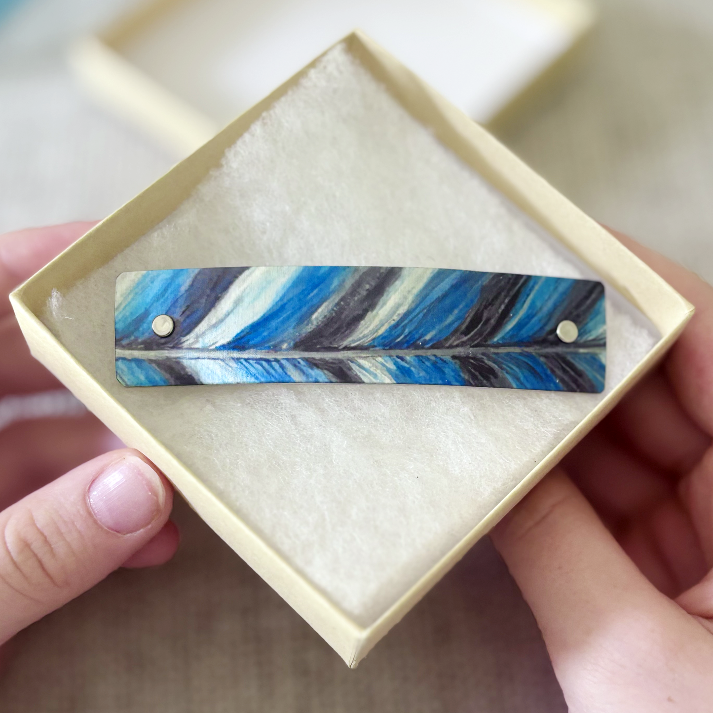 A handmade barrette hair clip featuring a blue, black, and white feather design, displayed inside a cream-colored square gift box held by hands.