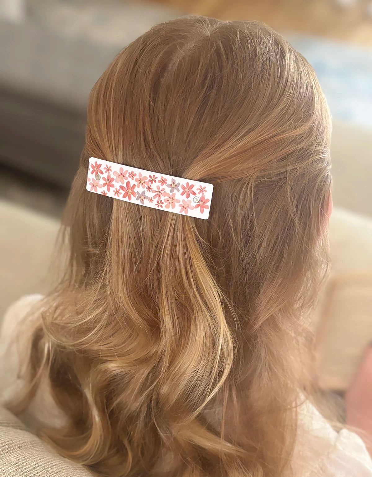 A close-up back view of a person with long blonde wavy hair, holding a white rectangular hair clip decorated with a pink and coral floral pattern.