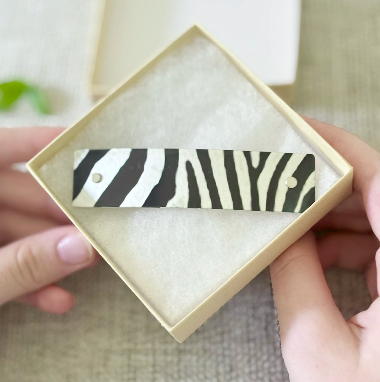 A zebra-print rectangular barrette with two small metal rivets, displayed inside an open cream-colored square gift box held by two hands.