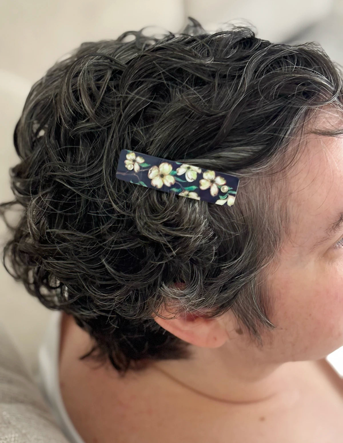 A close-up side view of a person with short, curly dark hair wearing a small rectangular hair clip decorated with artistic white dogwood flowers on a dark background.