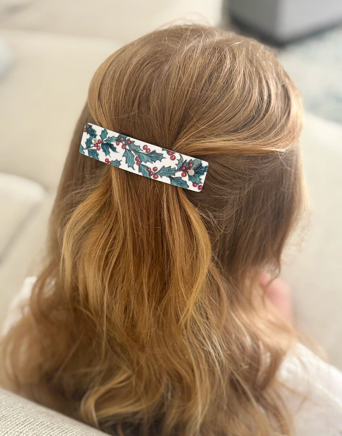 A close-up lifestyle shot of blonde hair from behind, secured with a rectangular hair clip decorated with a holly pattern on a white background.