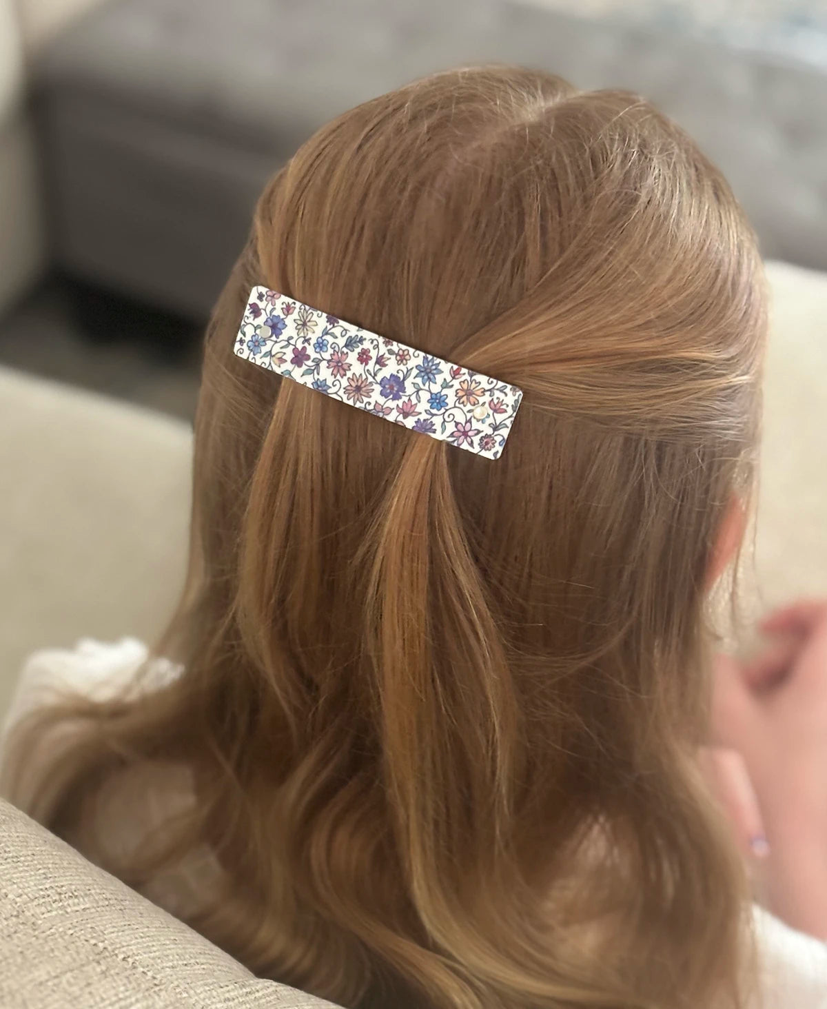 A close-up back view of a person with light brown hair wearing a slim rectangular hair clip adorned with a colorful floral pattern on a white background.