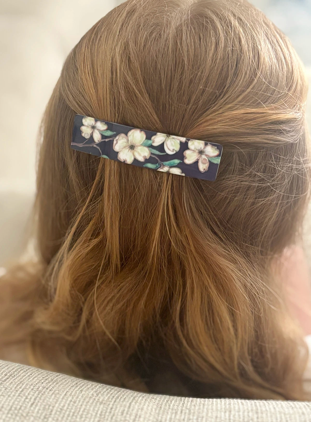 A close-up back view of a person with blond hair held by a  long rectangular hair clip. The clip has a charcoal gray background with a white floral dogwood pattern.