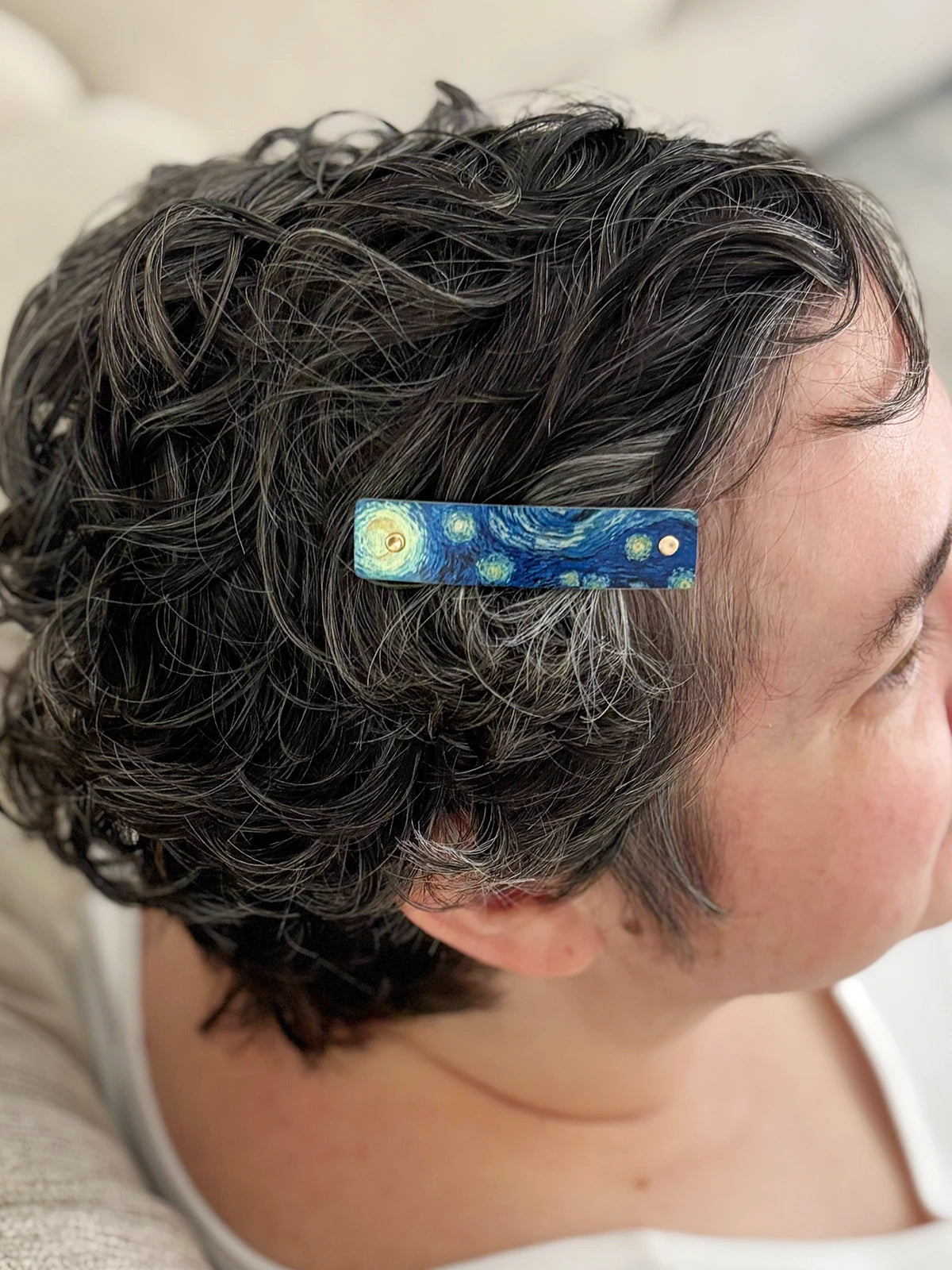 A close-up of a person with short, curly gray hair wearing a small bar-shaped hair clip decorated with a blue and yellow Starry Night style pattern.