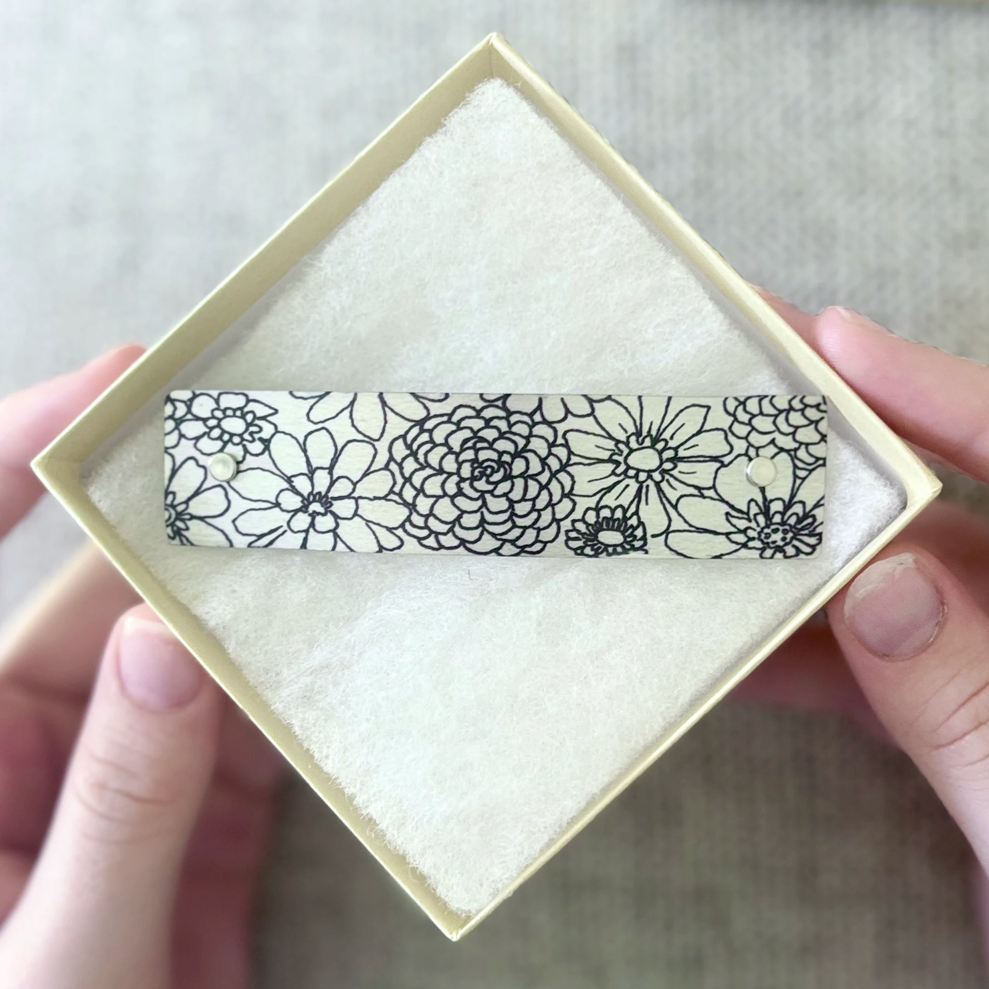 A close-up of a hand holding an open cream-colored jewelry box containing a silver hair barrette decorated with ink drawn zinnia flowers.