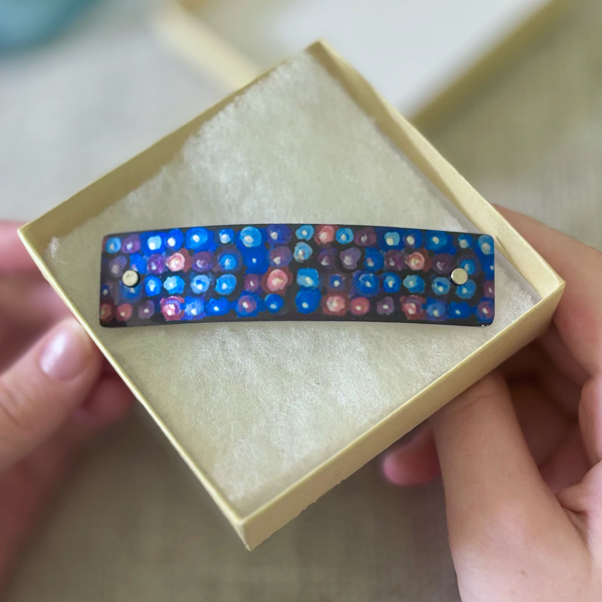 A handmade rectangular barrette hair clip with a blue and multicolor blackberry pattern, presented in a small beige jewelry box held by hands.