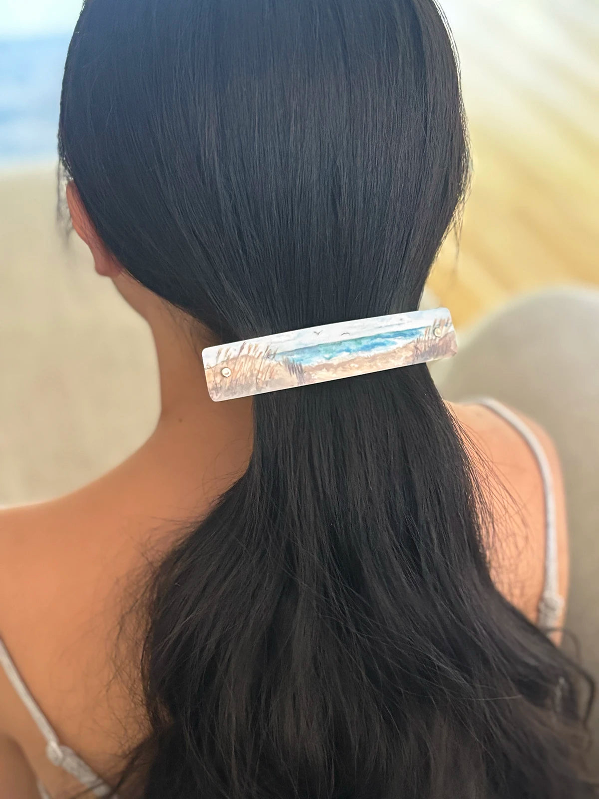 A woman viewed from behind with long black hair, wearing a rectangular hair clip featuring a watercolor beach scene with blue water and beige sand.