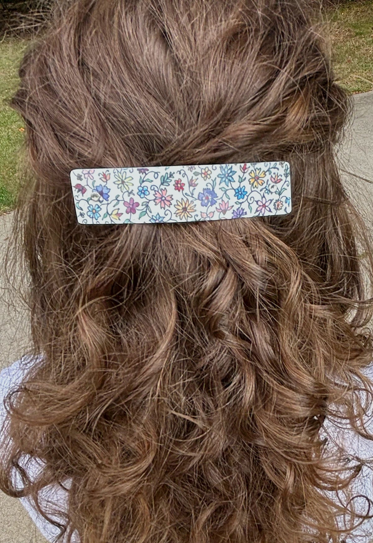 A close-up back view of a person with long, wavy brown hair wearing a rectangular hair clip adorned with a colorful floral pattern on a white background.