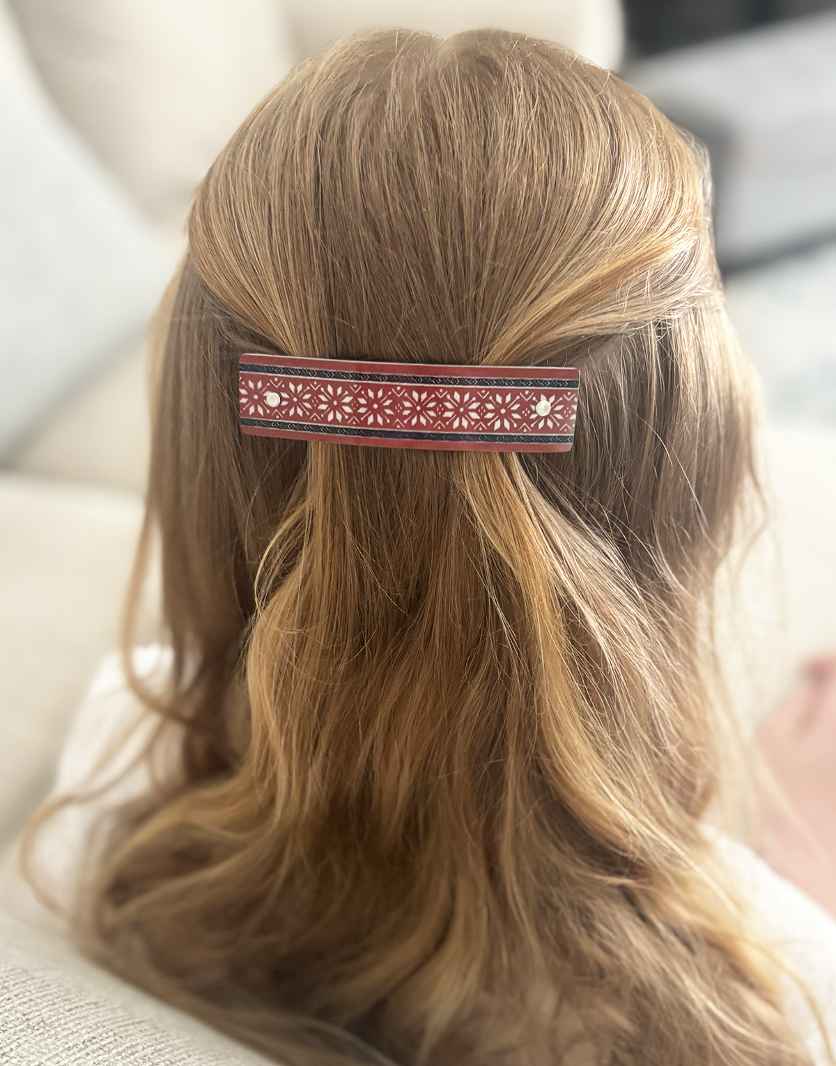 A close-up of a person's blonde hair secured with a decorative hair clip. The clip is a narrow horizontal bar with a red background and a repeating white floral or geometric pattern, bordered by thin black lines.