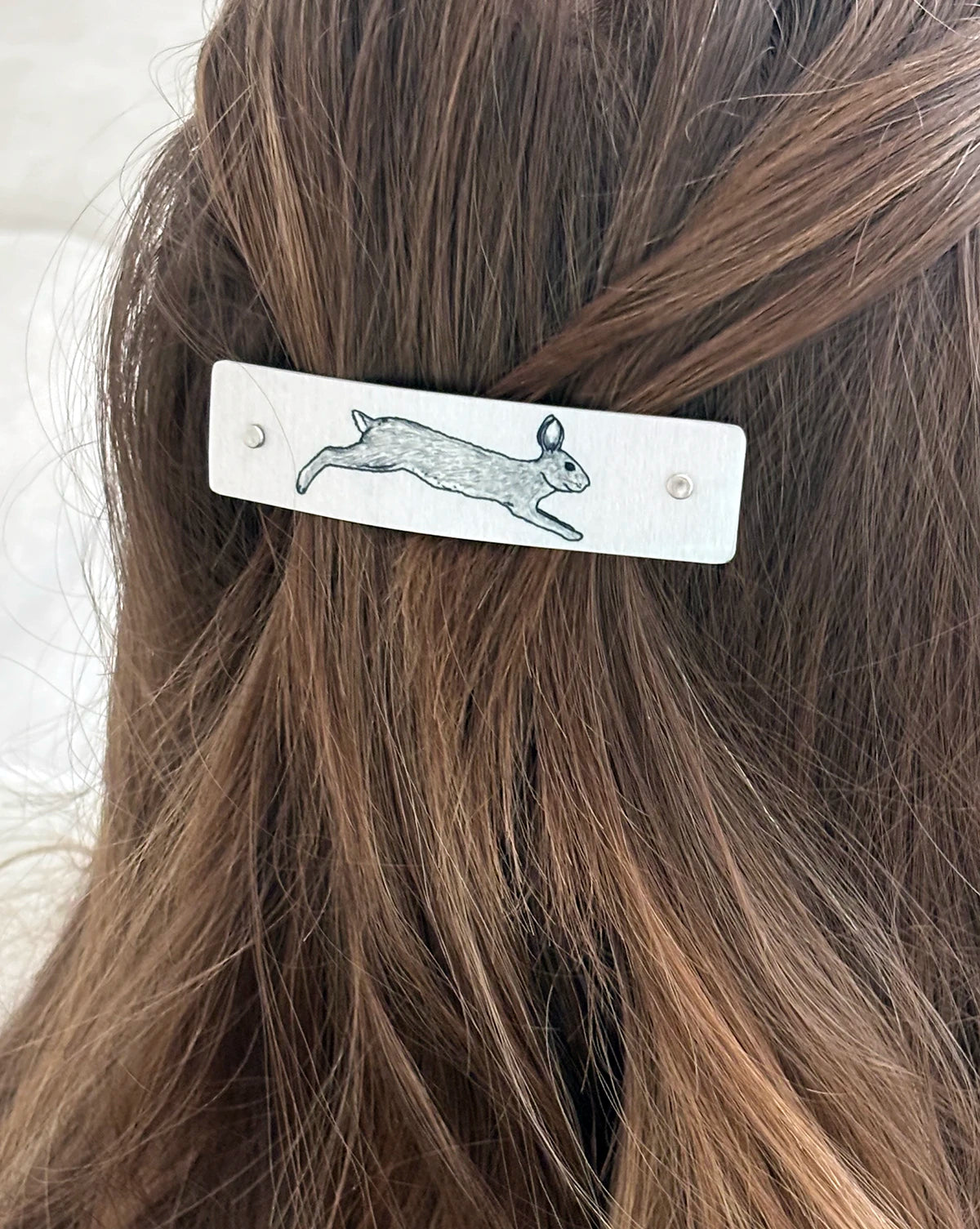 A close-up of a person's brown hair secured with a silver rectangular hair clip featuring an original black ink drawing of a leaping rabbit.