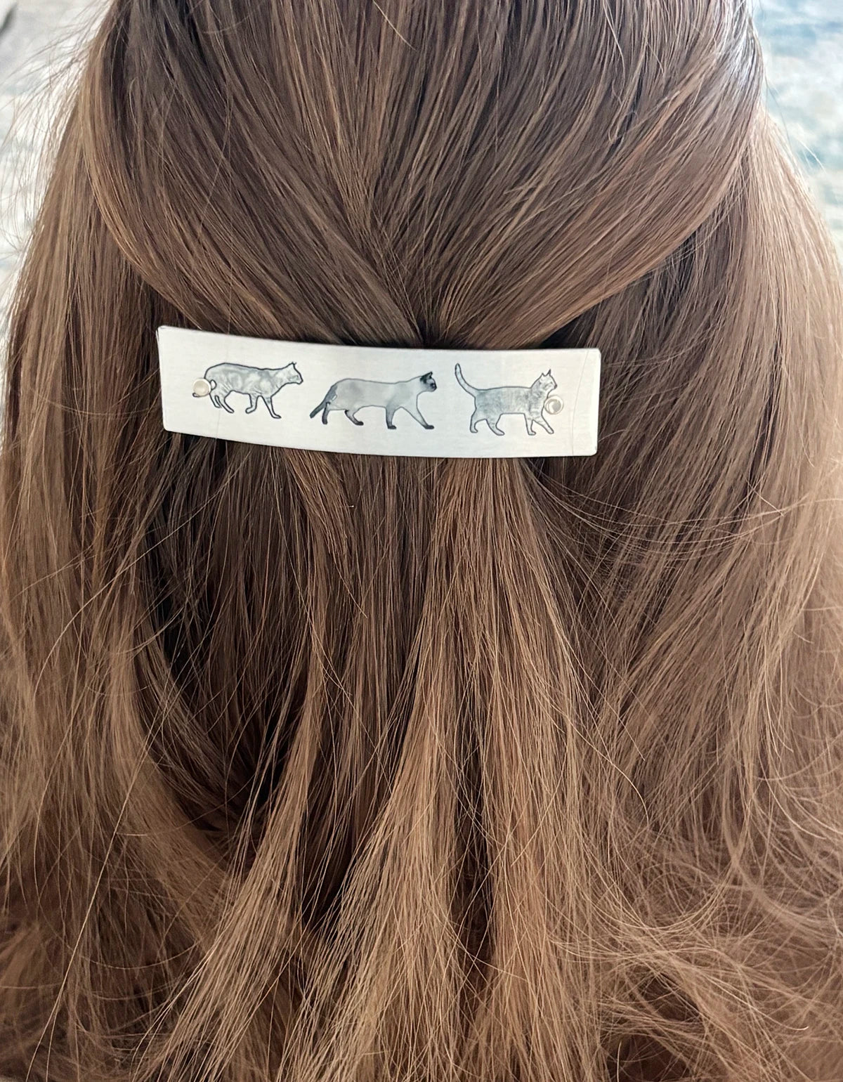 A close-up of a person's brown hair featuring a silver rectangular hair clip decorated with a row of walking cat illustrations.