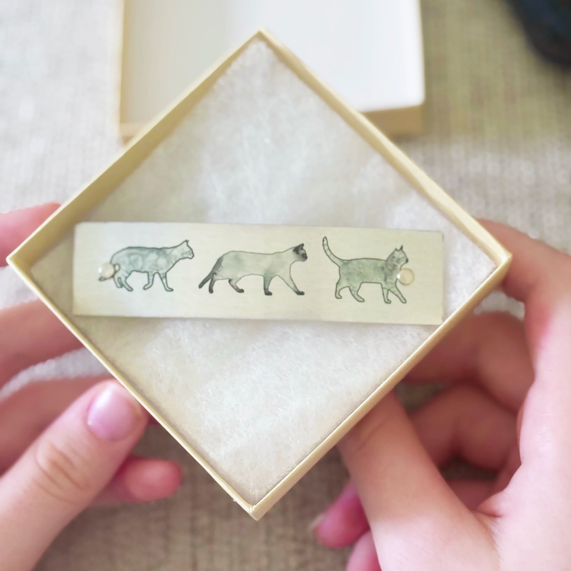 A close-up of hands holding an open square cream-colored gift box with a boutique hair clip inside. The barrette features three illustrated cats in muted gray and black tones on a silvery background.