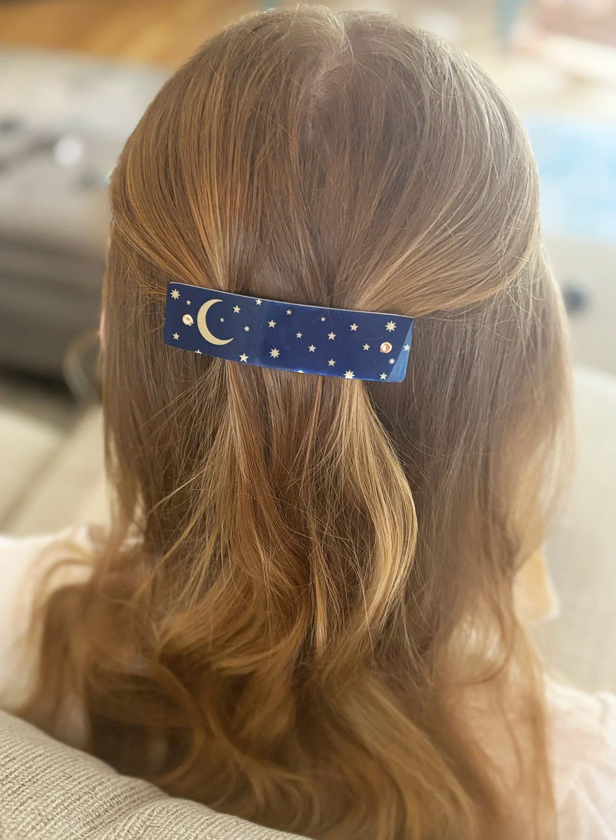 A close-up back view of a person with strawberry blond hair wearing a navy blue hair clip decorated with a gold crescent moon and stars.