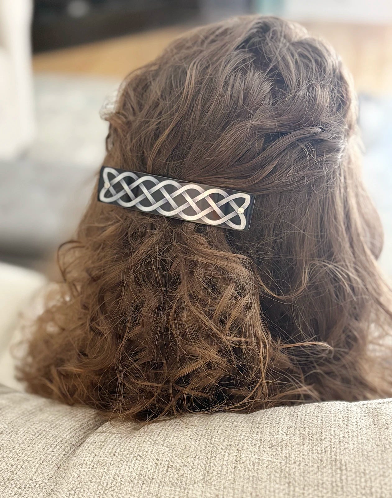 A close-up back view of a person with brown curly hair wearing a black and silver metal hair clip with an intricate Celtic knot design.